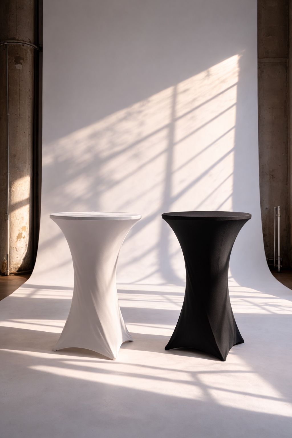Zwei Tall-Tables, eine weiße und eine schwarze, vor einem weißen Hintergrund mit Schatten durch Fenster. Traditioneller Industriebau mit sichtbaren Wänden auf der linken und rechten Seite.