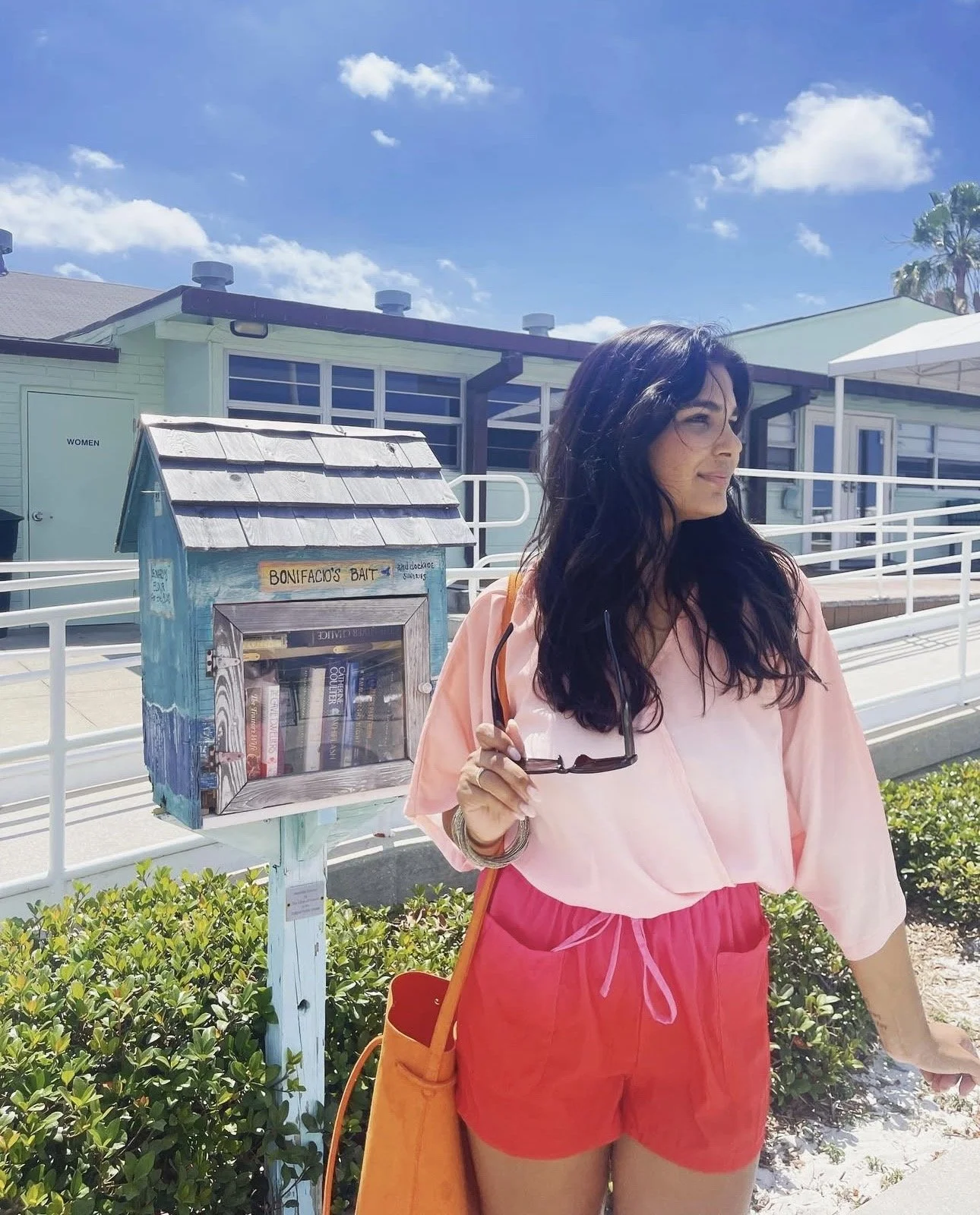 Woman outdoors holding sunglasses, wearing pink and red outfit, standing near a blue beach library box with a sign 'Bonificio's Bait,' on a sunny day with blue sky and clouds. Vania