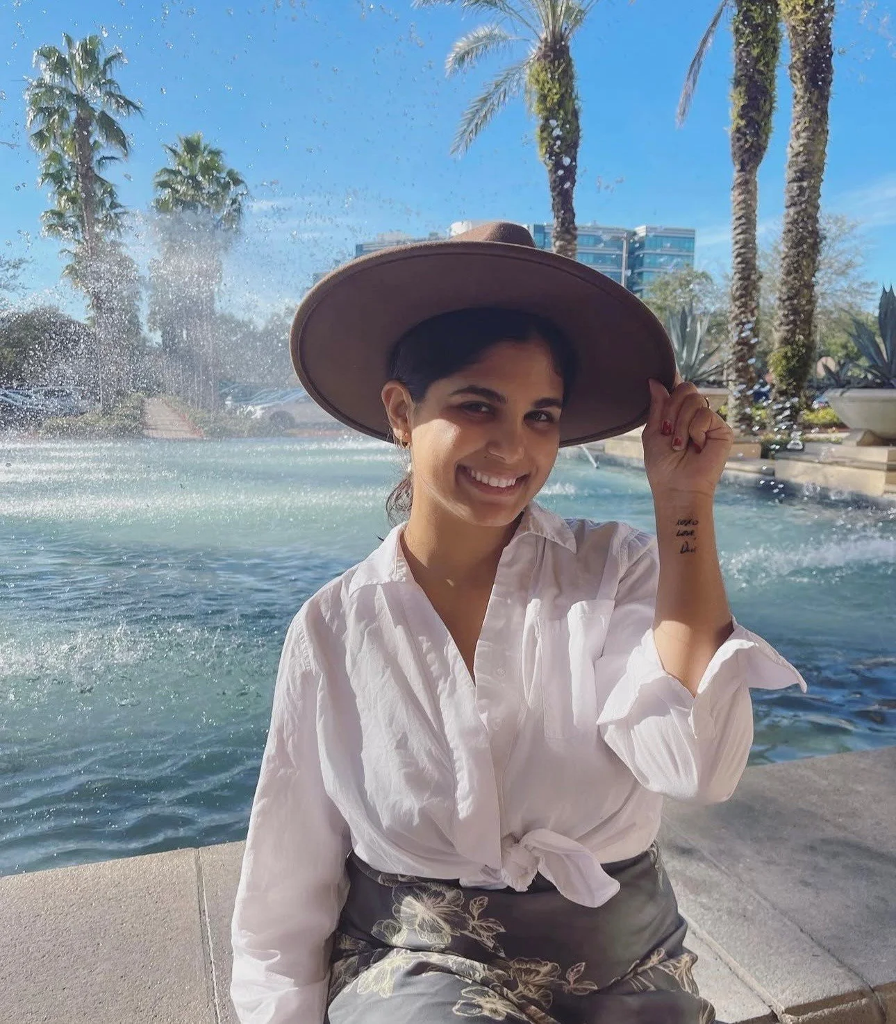 A woman smiling and wearing a large hat, white blouse, and floral skirt, sitting near a fountain with palm trees and buildings in the background on a sunny day. Vania
