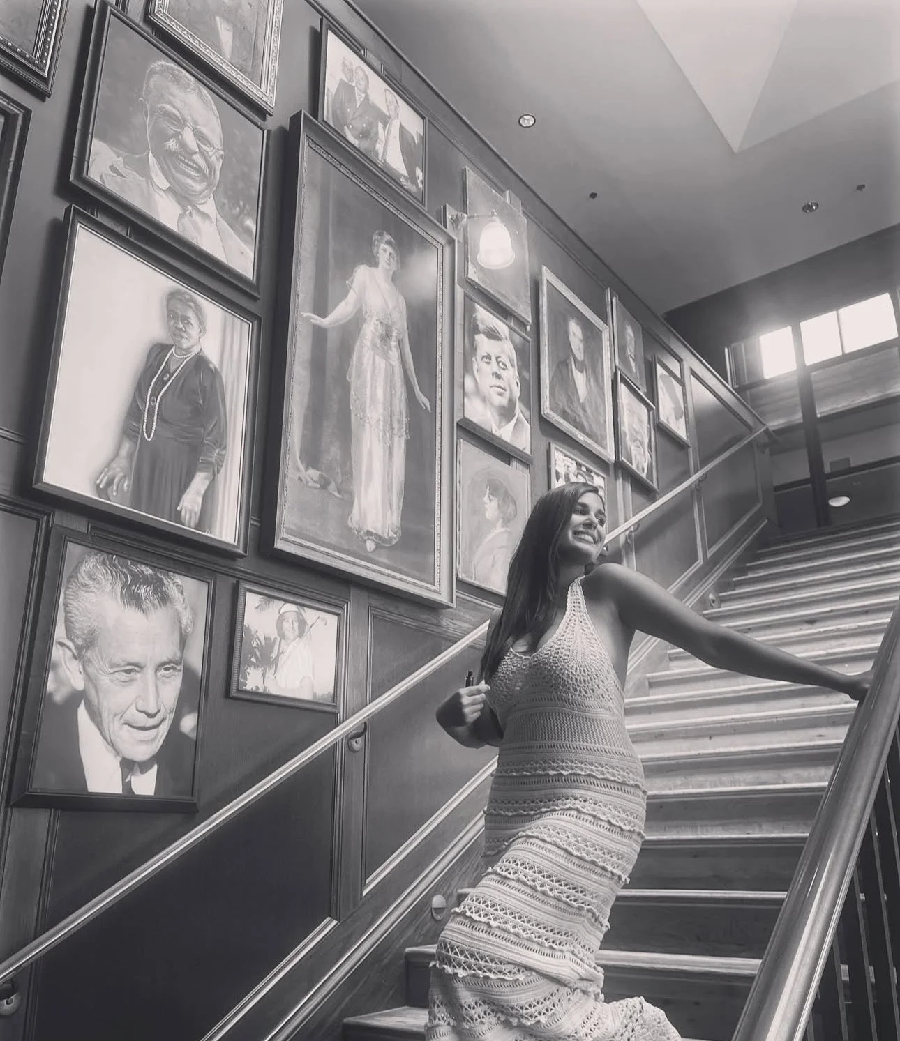 A woman in a crochet dress smiling and posing on a staircase with a wall of framed black-and-white portraits in the background. Vania