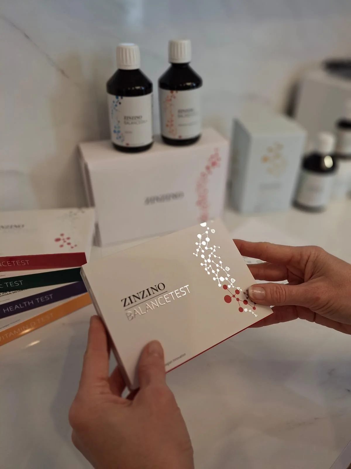 A person's hand holds a box labeled "ZINZINO BALANCE TEST" with a molecular design graphic. In the background, there are medical bottles, boxes, and test kits on a white surface.
