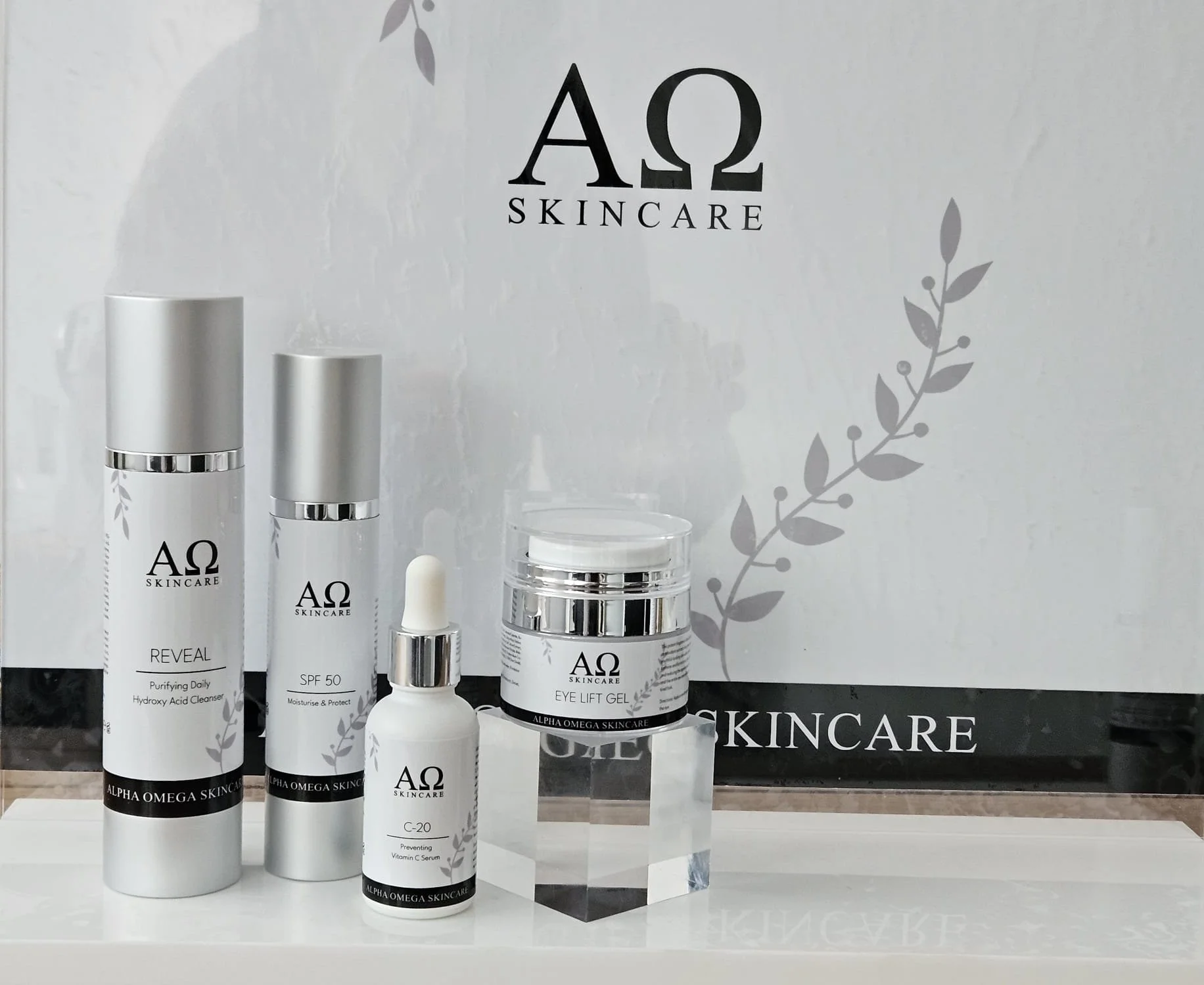 Skincare products from Alpha Omega Skincare, including a purifying daily hydroxy acid cleanser, SPF 50 moisturizer, vitamin C serum, and eye lift gel, displayed in front of a branded backdrop.