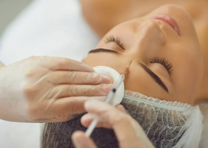 Close-up of woman receiving a facial treatment, with her eyes closed, lying down with a hair cover, as a skincare professional applies a product with a cotton pad.