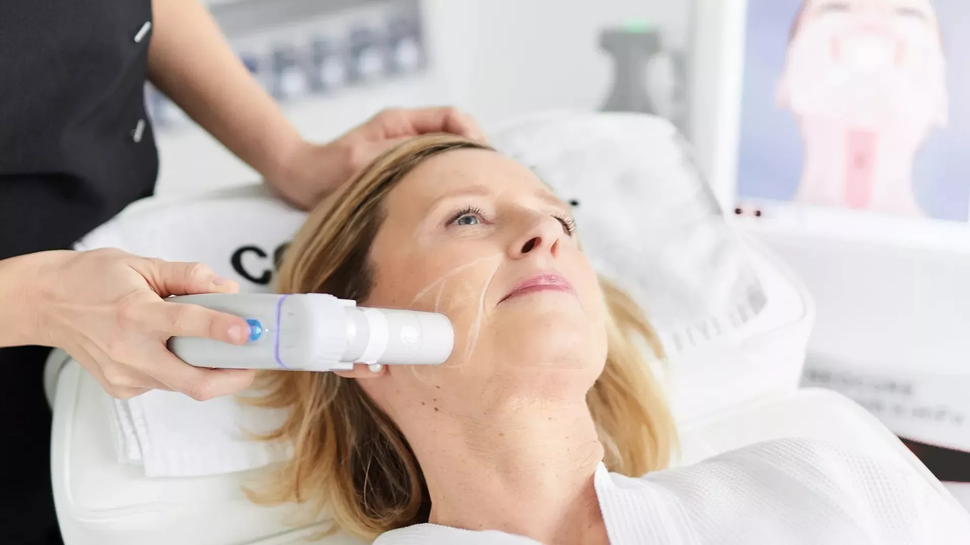 A woman receiving a cosmetic facial treatment with a device applied to her cheek in a clinical setting.