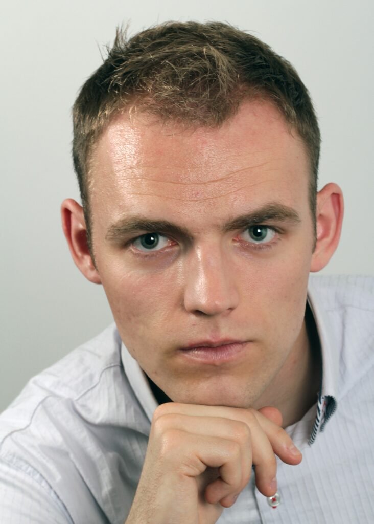 A young man with short, light brown hair and blue eyes looking directly into the camera, resting his chin on his hand, wearing a white collared shirt against a plain background.