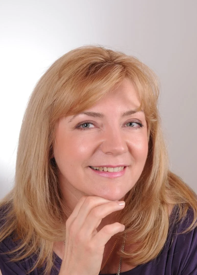 A woman with blonde hair smiling and resting her chin on her hand, wearing a purple top.