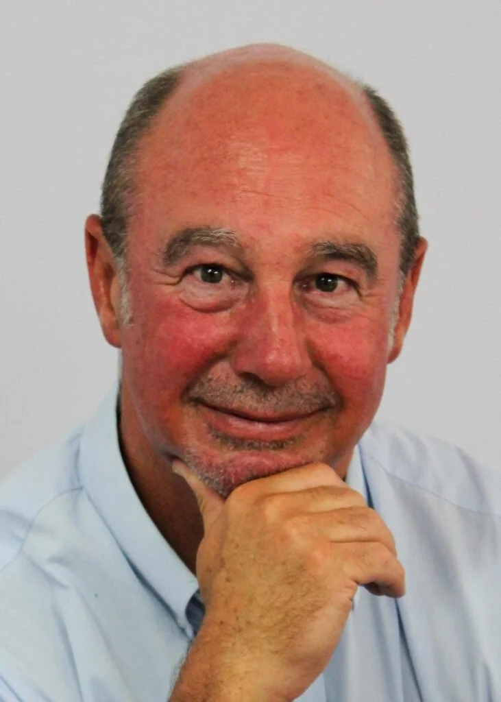 A close-up portrait of a middle-aged man with a bald head and a slight beard, smiling and resting his chin on his hand, wearing a light blue shirt.
