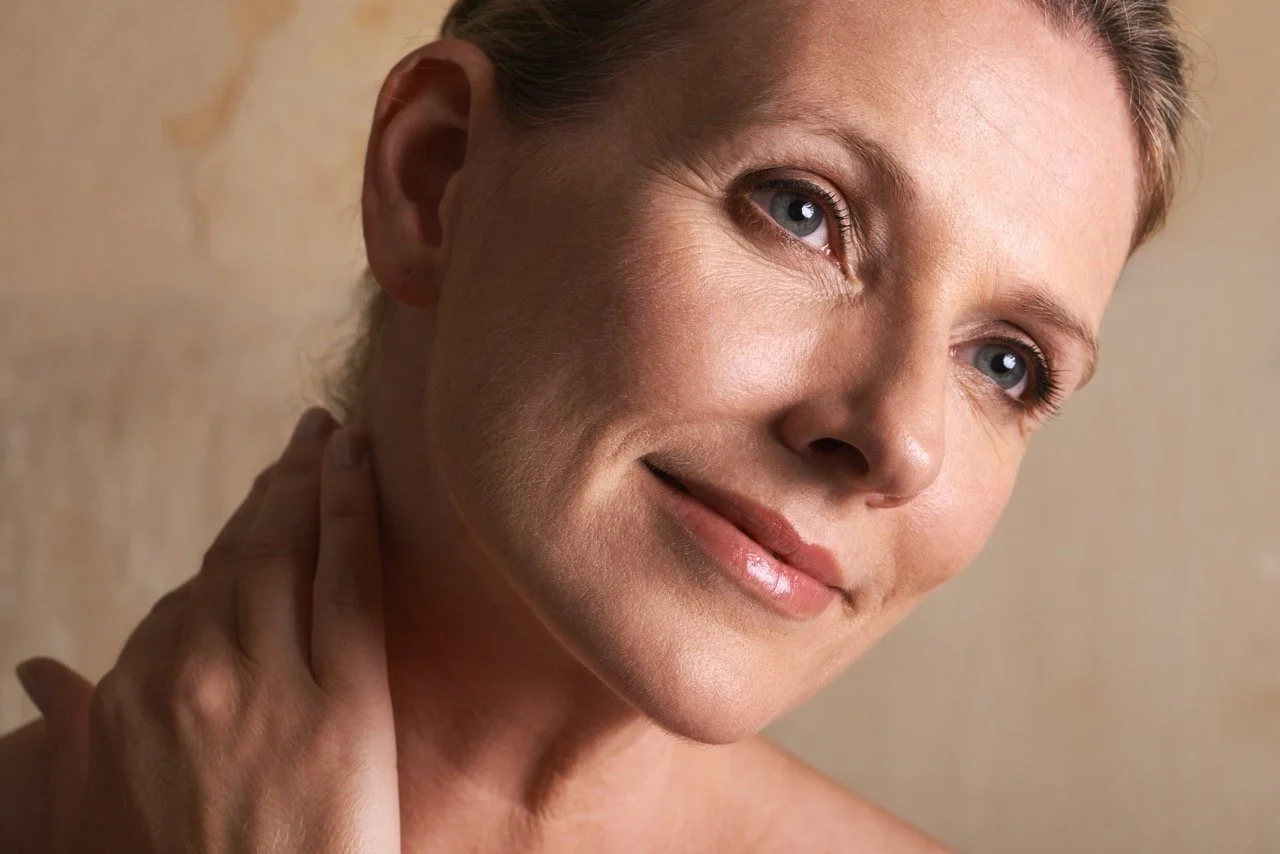 A close-up of a middle-aged woman with short hair, smiling gently, touching her neck with her hand, against a neutral background.