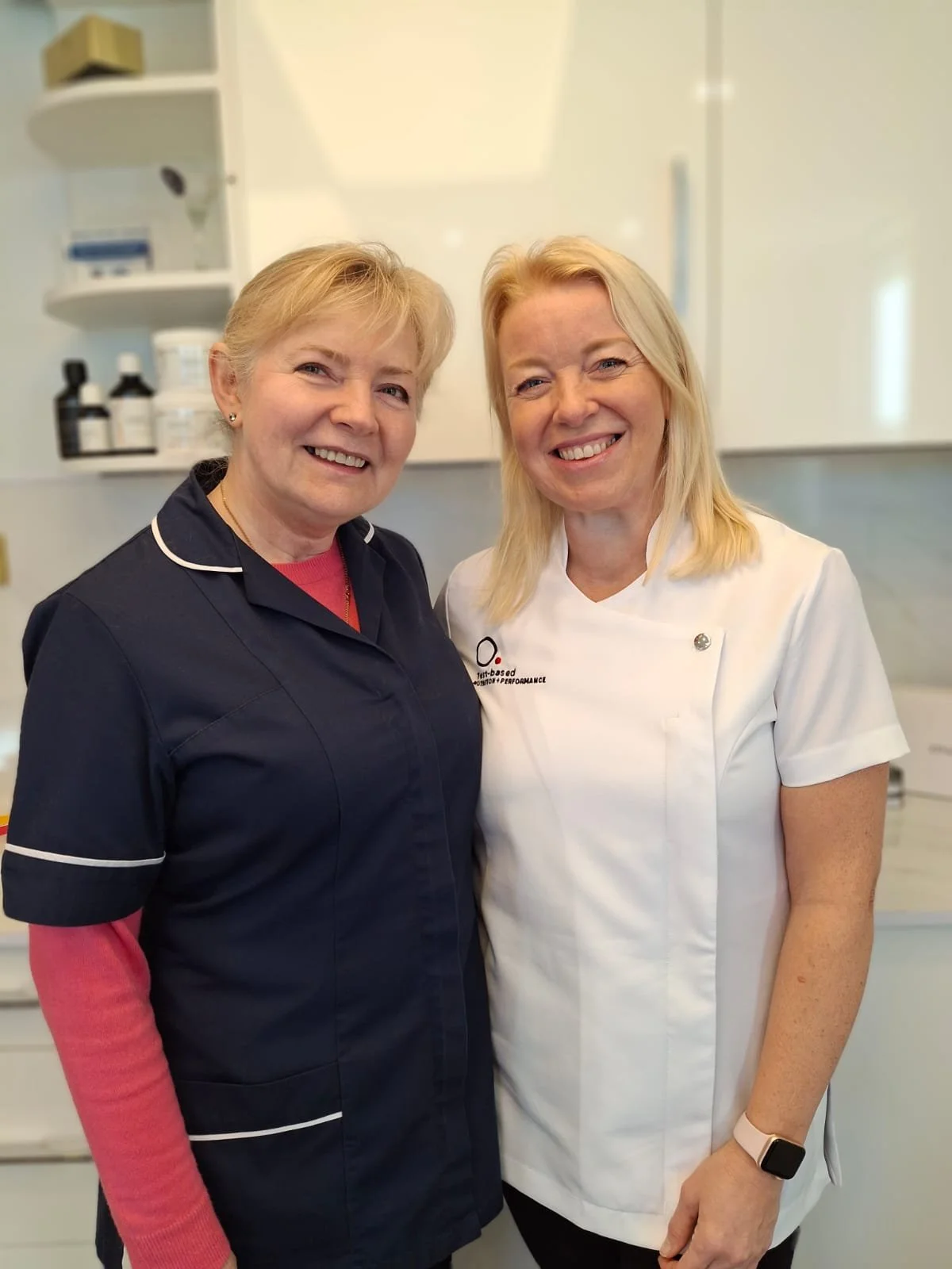 Two women, one in navy scrubs and the other in a white medical coat, are smiling and standing close together in a clinical setting with medical supplies on shelves behind them.