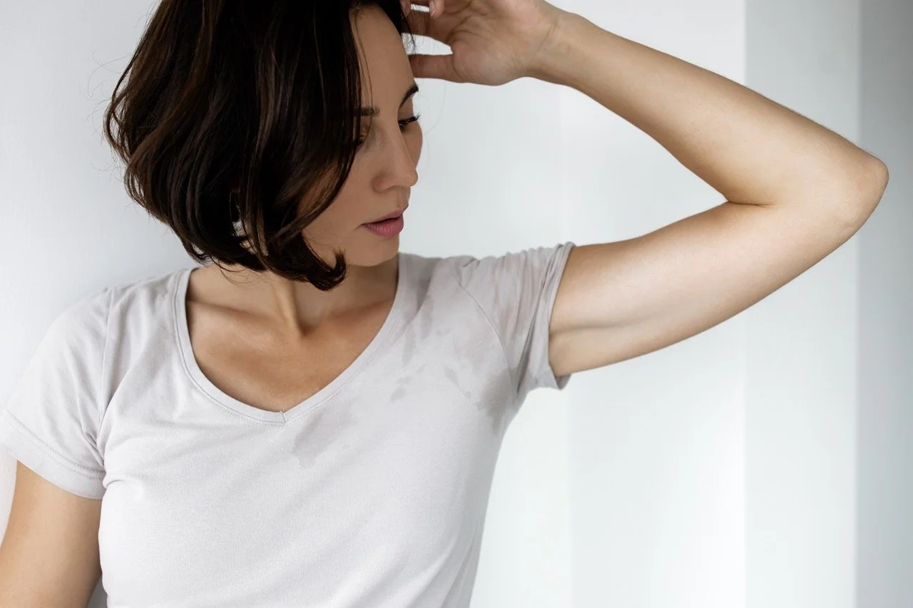A woman with shoulder-length dark hair in a white t-shirt touching her forehead while looking down, standing against a plain white wall.
