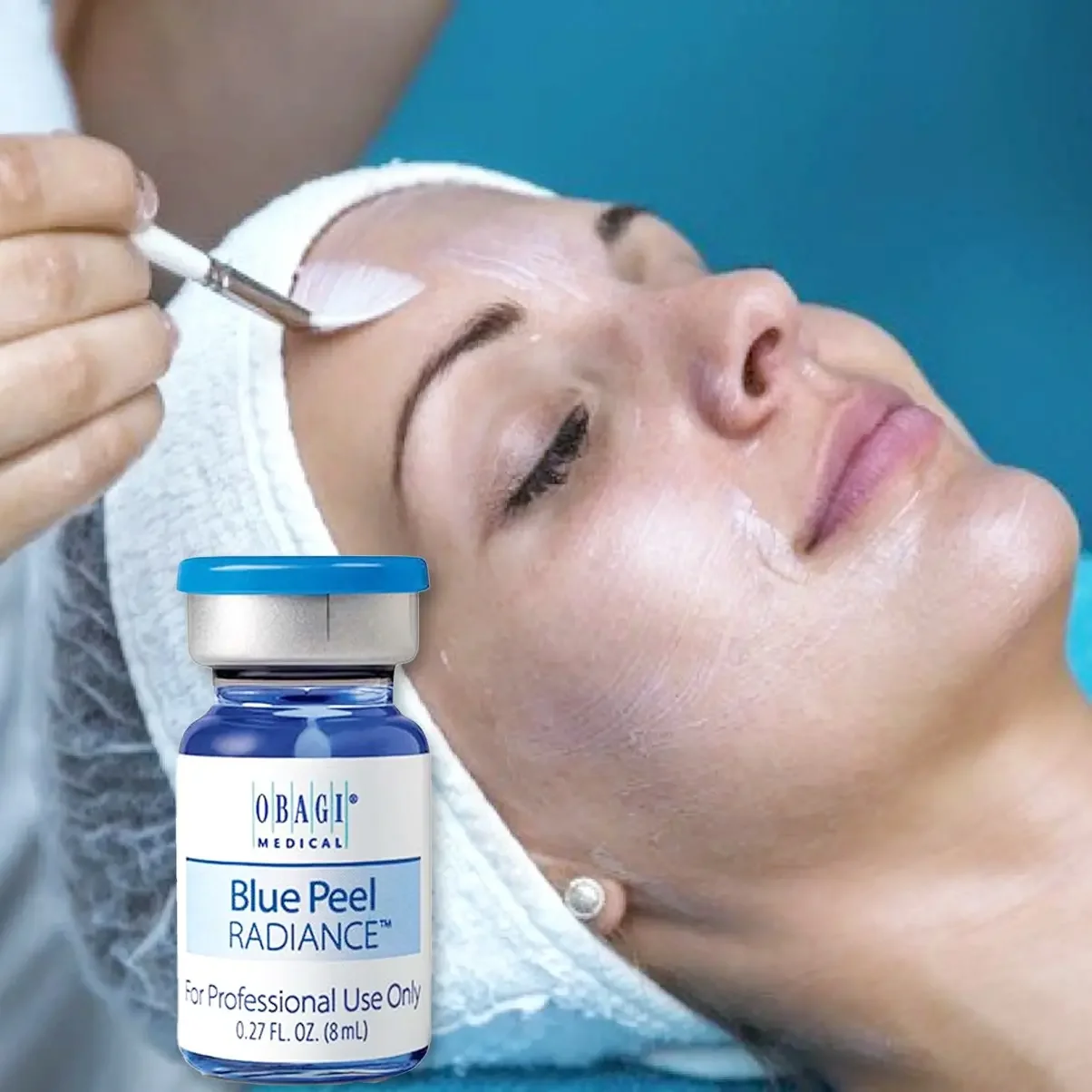 A woman lying on a spa bed receiving a facial treatment with a dropper applying a liquid to her forehead. There is a bottle of Obagi Blue Peel Radiance product in the foreground.