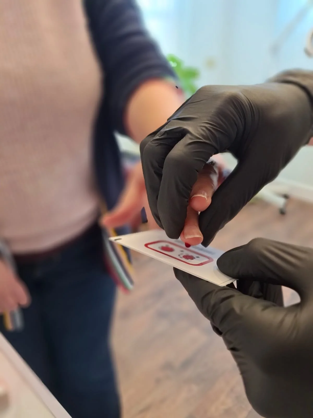 Person receiving a blood test, with a healthcare professional using black gloves to prick a finger.