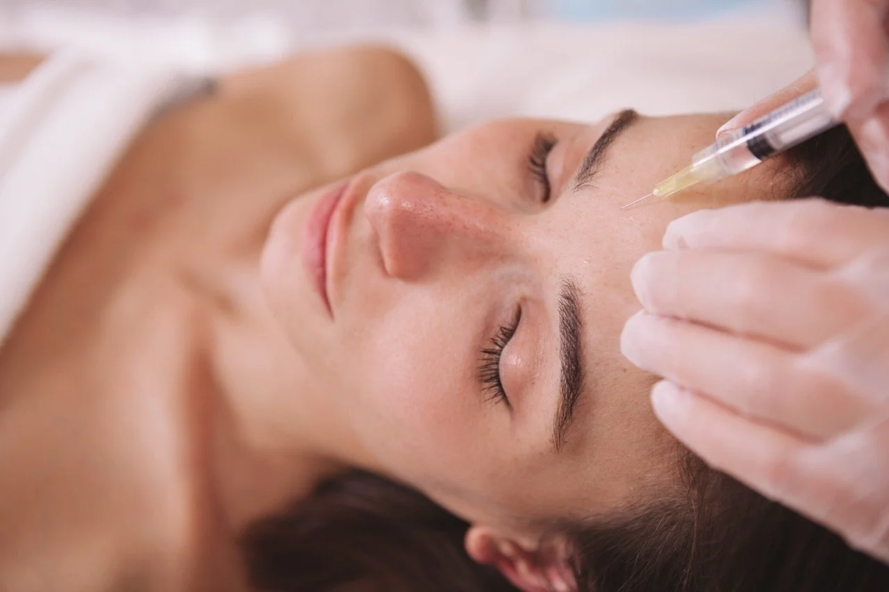 A woman receiving a cosmetic injection in her forehead by a medical professional wearing gloves.