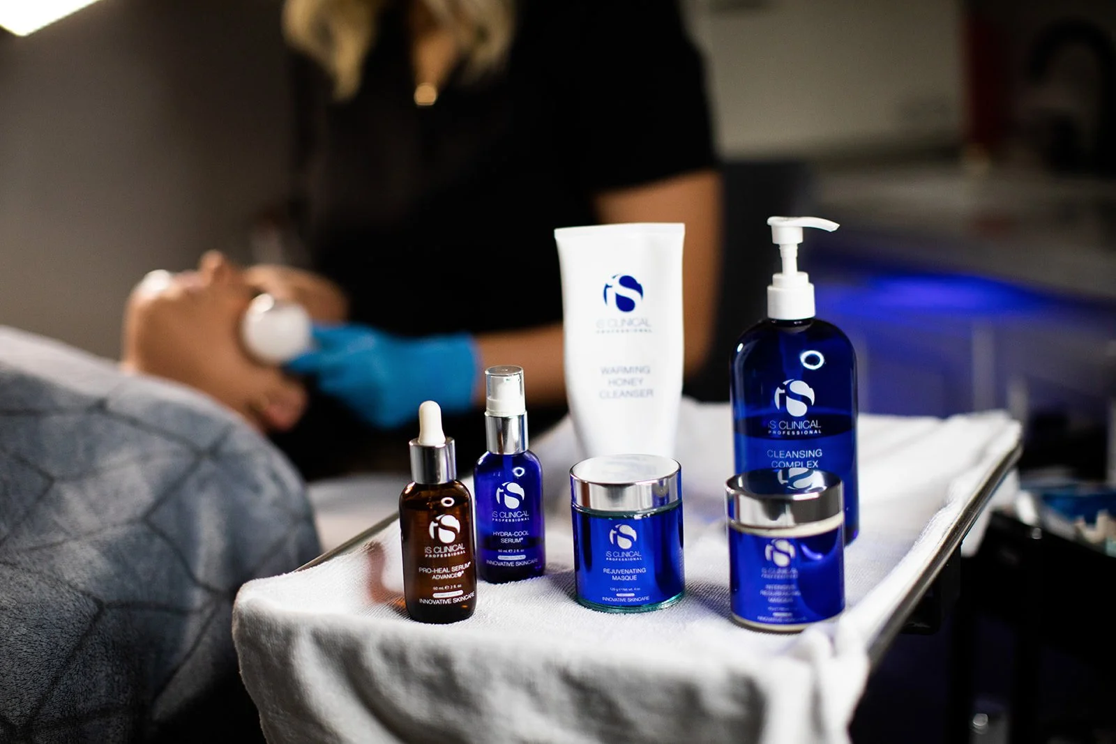 A person wearing black clothing and blue gloves performing a facial treatment on a client lying back with eyes closed, while various skincare bottles and jars are arranged on a white towel in the foreground.