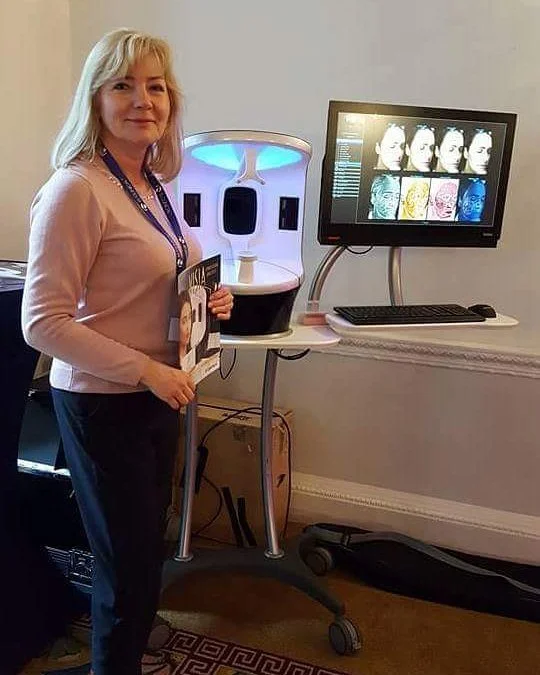 A woman standing next to a skincare analysis machine with a computer monitor displaying various facial images and skin analysis data.