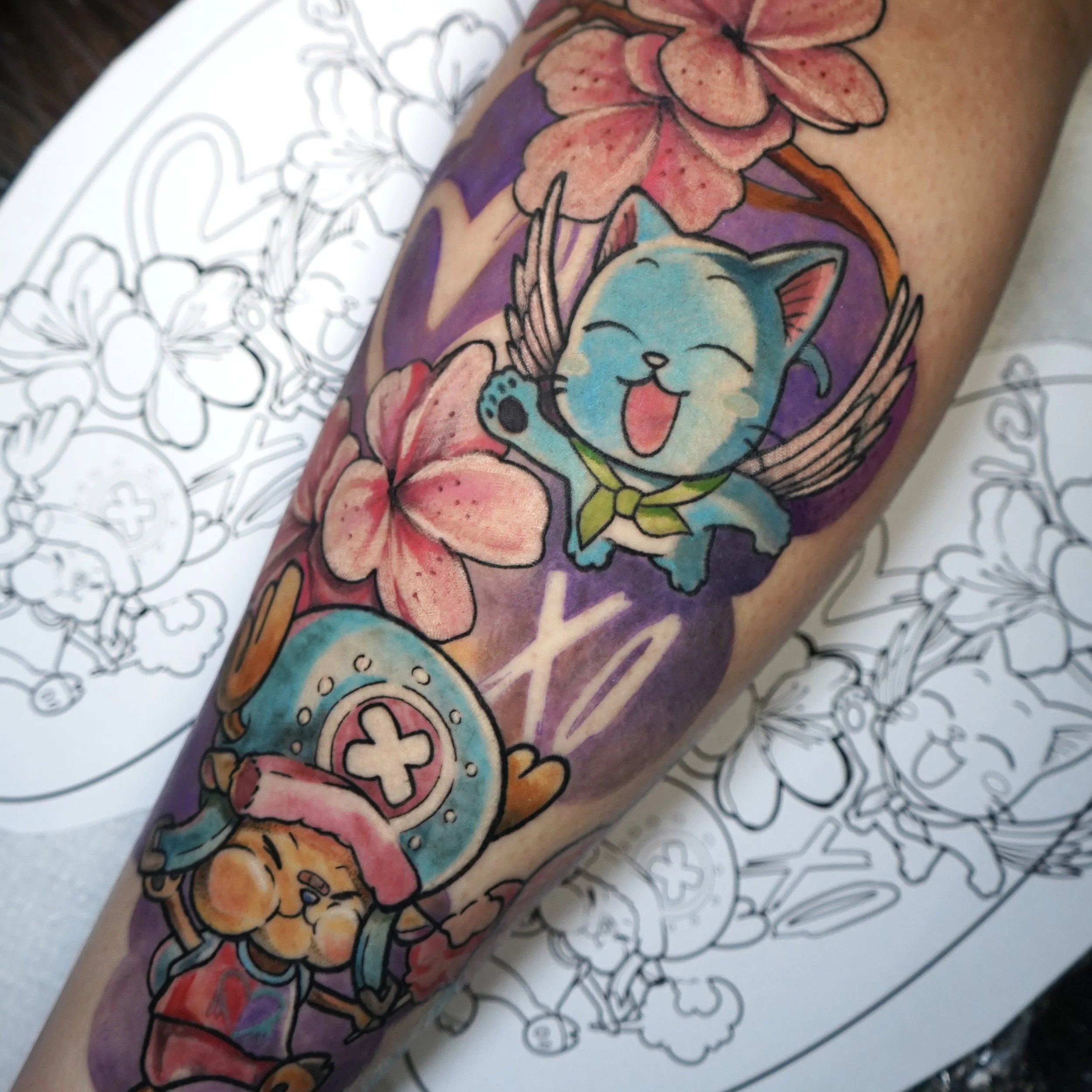 Colorful tattoo of a cute, blue cartoon cat with fairy wings and a yellow neckerchief, surrounded by pink flowers, on a person's arm. A black and white outline of the same design is visible on paper beneath.
