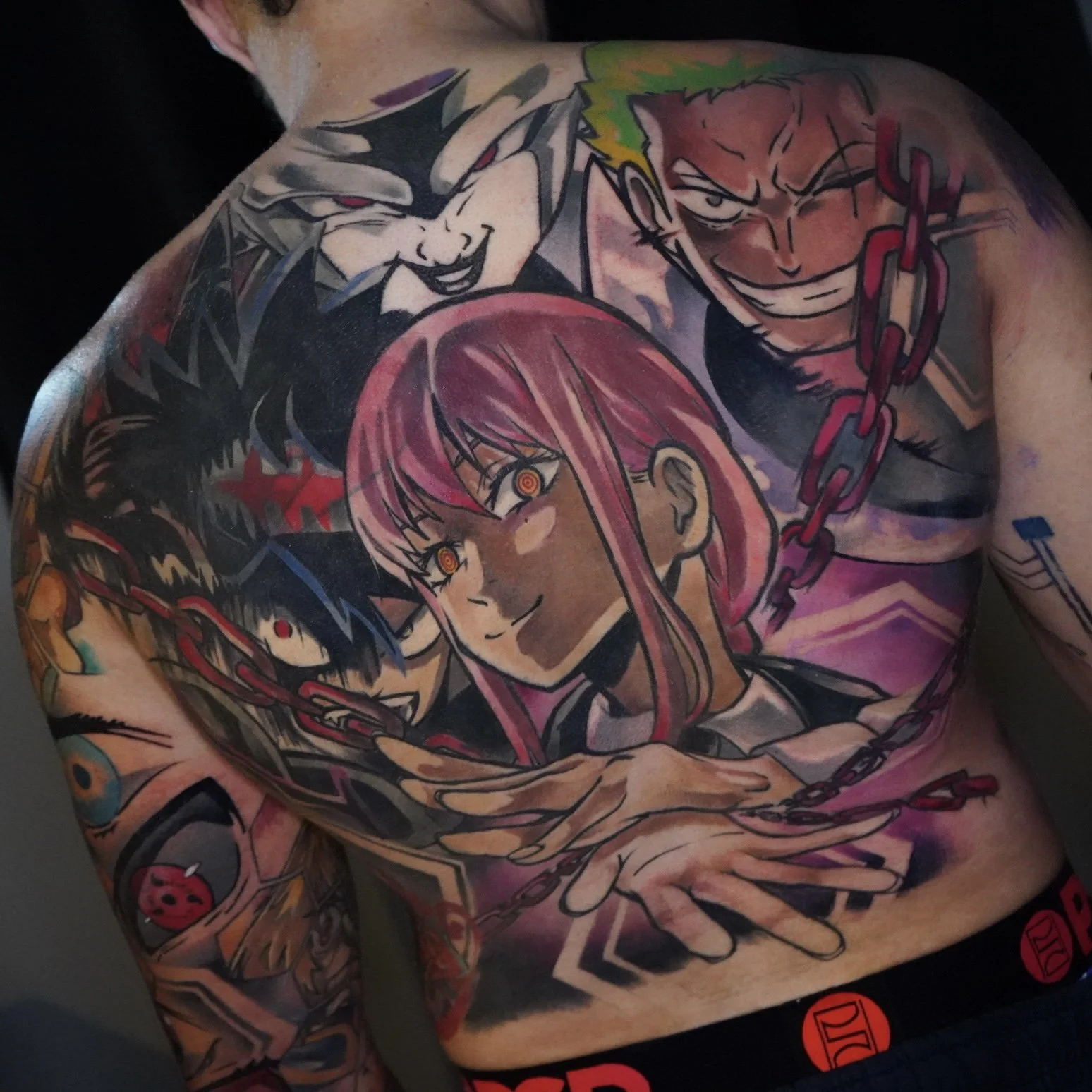 Colorful tattoo of three anime characters on a person's chest and shoulder, featuring a woman with pink hair and yellow eyes, a man with spiky green hair and a mischievous grin, and a dark-haired boy with a headband, all connected by chains.