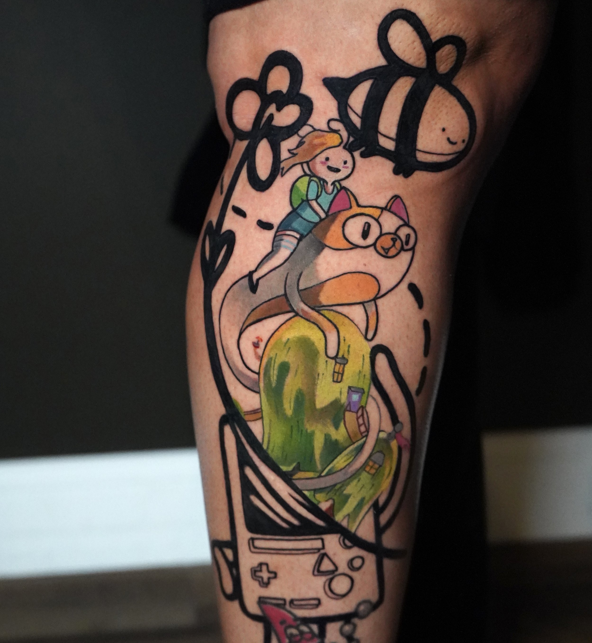 Colorful tattoo of a girl riding a cat with glasses, a bee, a tablet, a watermelon, and various doodles on the arm.