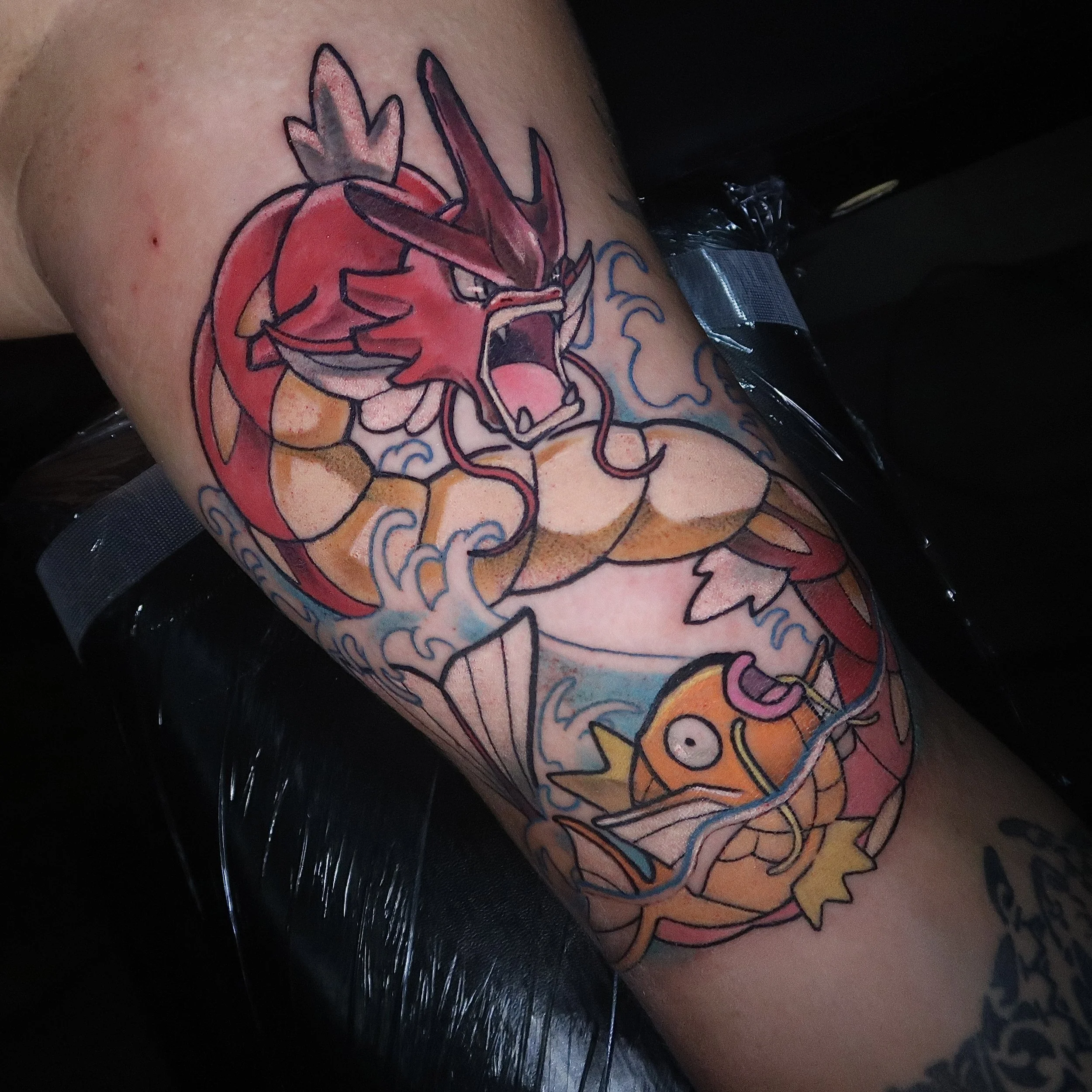 Tattoo of a red and yellow dragon with blue water waves and an orange fish with a surprised expression on a person's upper arm.