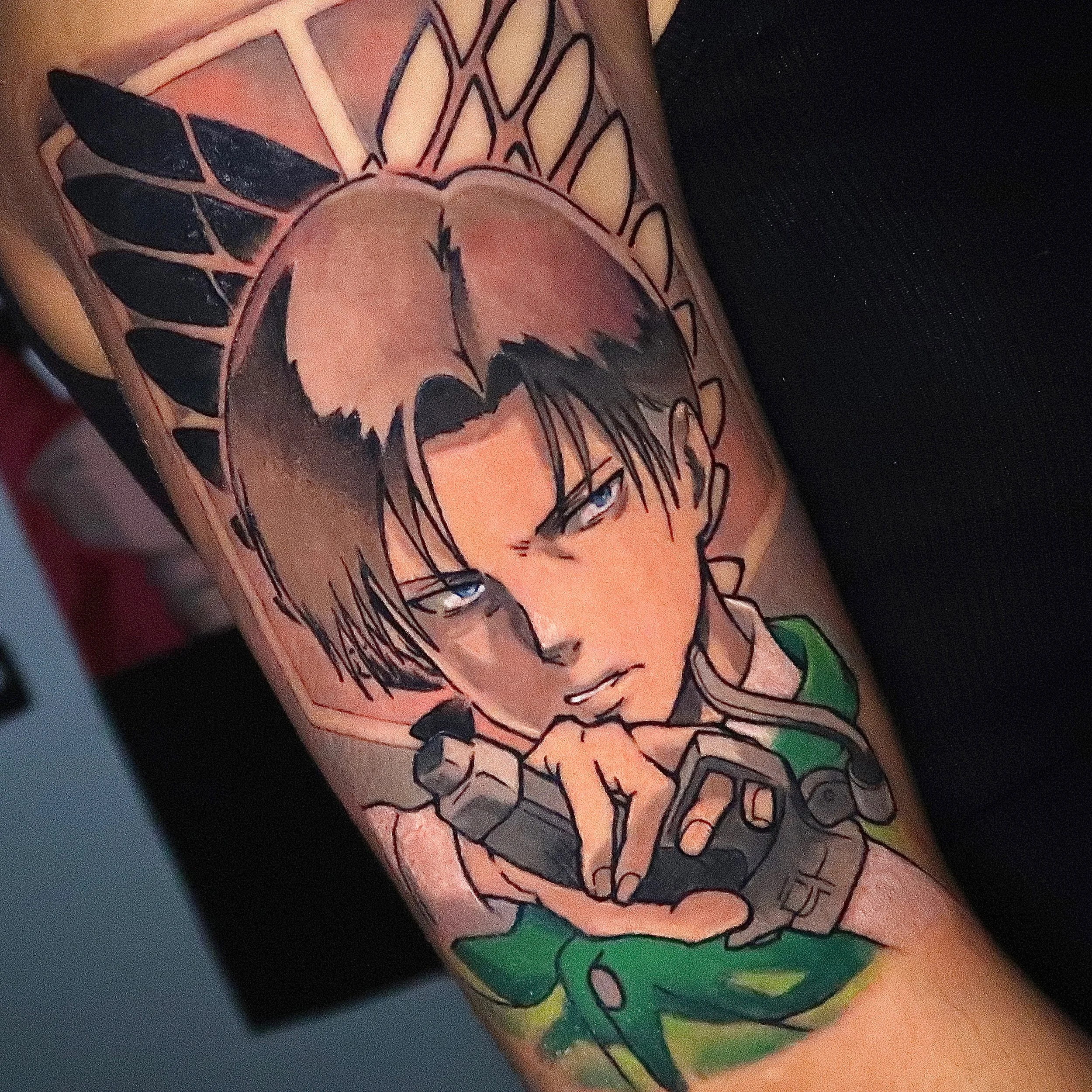 Tattoo of a cartoon boy with blue eyes, black hair, and a serious expression, wearing a green shirt and holding a game controller, with tribal-style black and orange feathers behind his head.