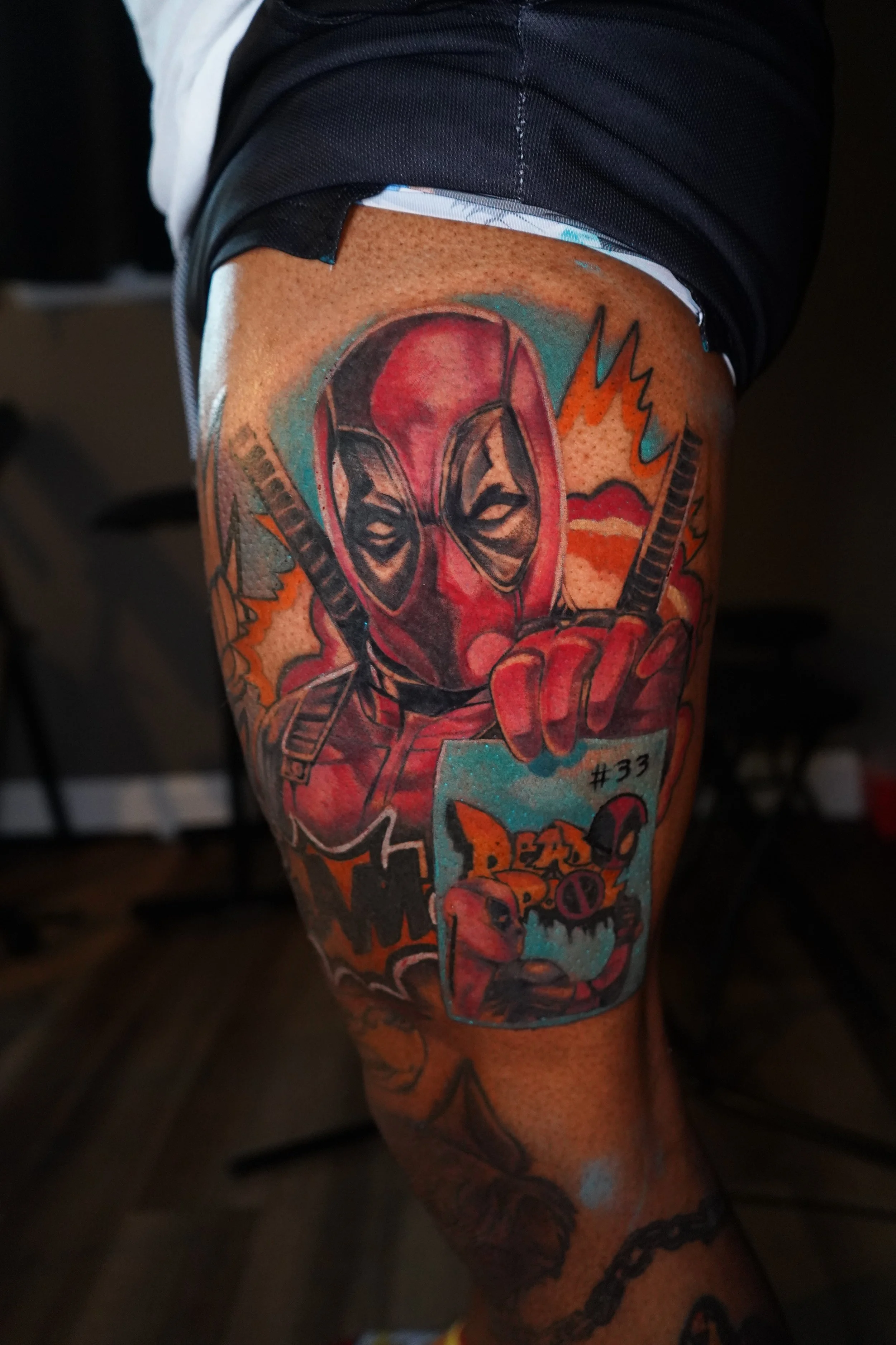Tattoo of Deadpool with a finger pointing outward, holding a card with a dog's face, and comic-style background elements on a person's thigh.