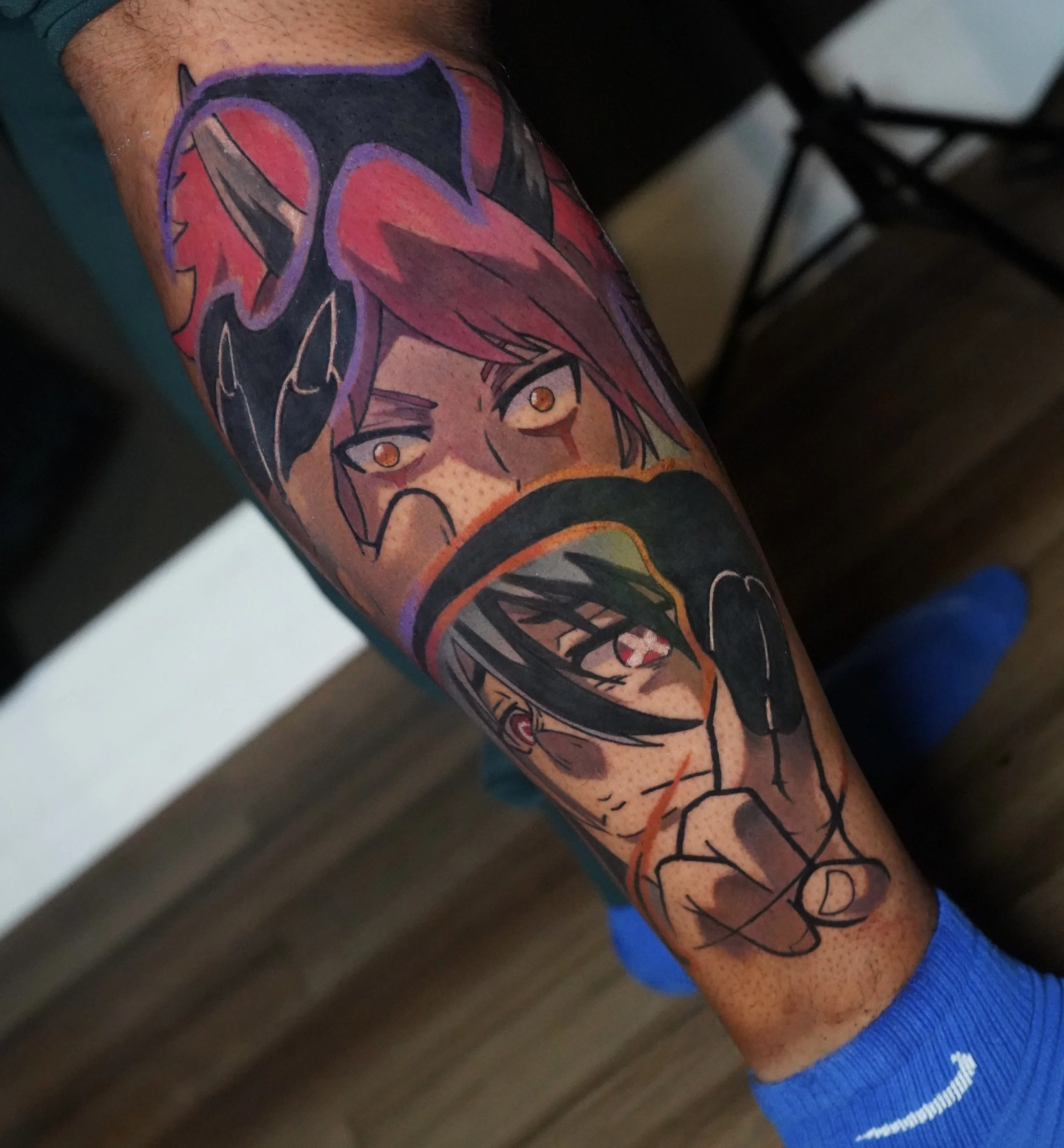 Tattoo of two anime-style characters with vibrant hair, one with red hair and the other with dark hair, and a hand making a shushing gesture.