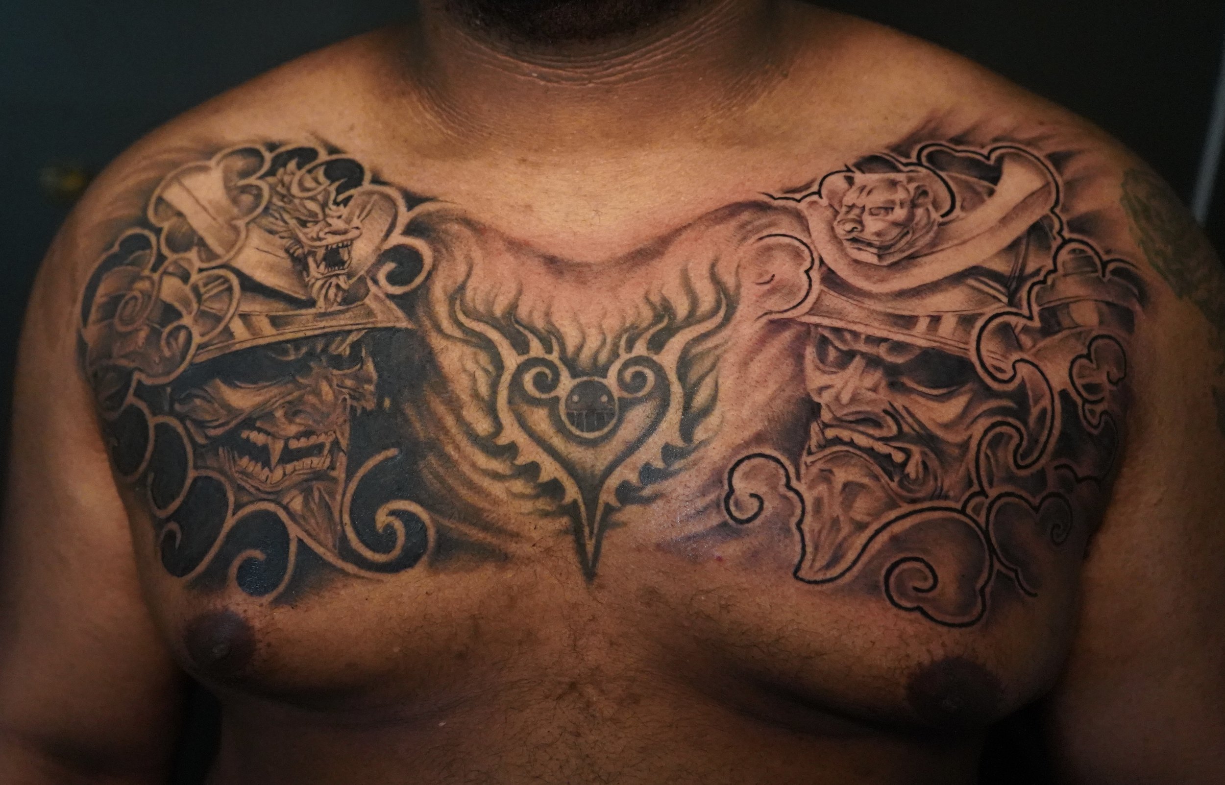 Tattoo of a heart with flames in the center on a man's chest, flanked by two fierce tiger and demon faces, with intricate swirling patterns.