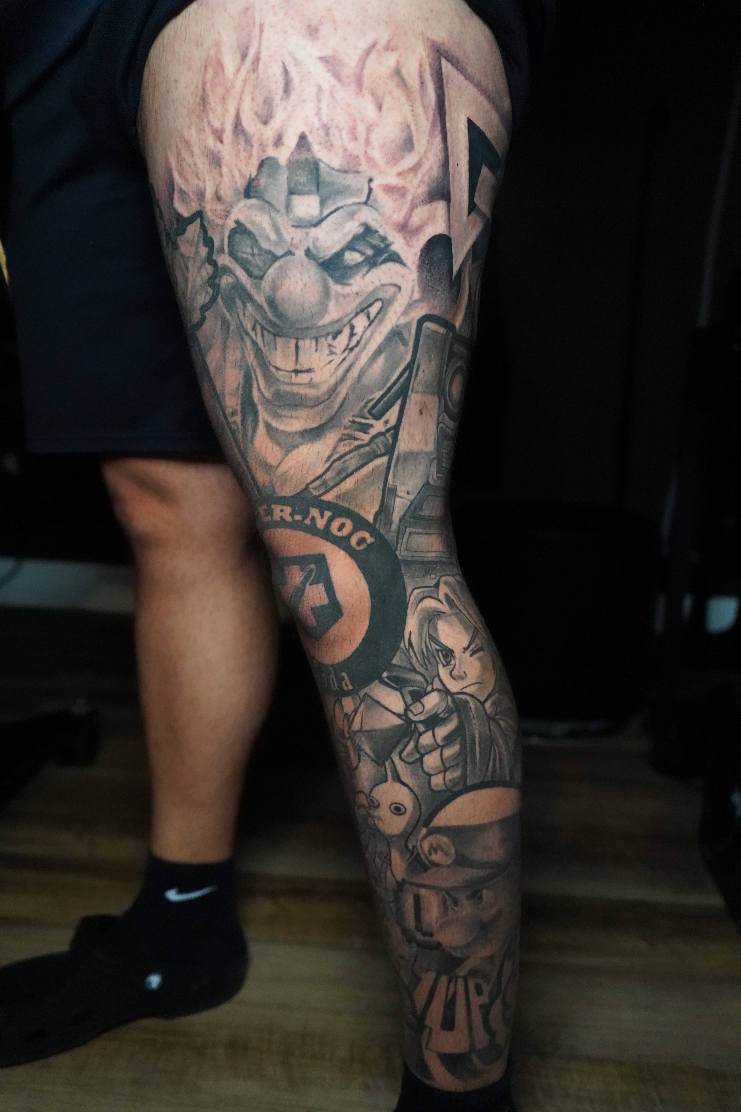 A detailed black and gray tattoo sleeve featuring characters from video games, including a large clown face, a character holding a gun, and other gaming-related imagery.