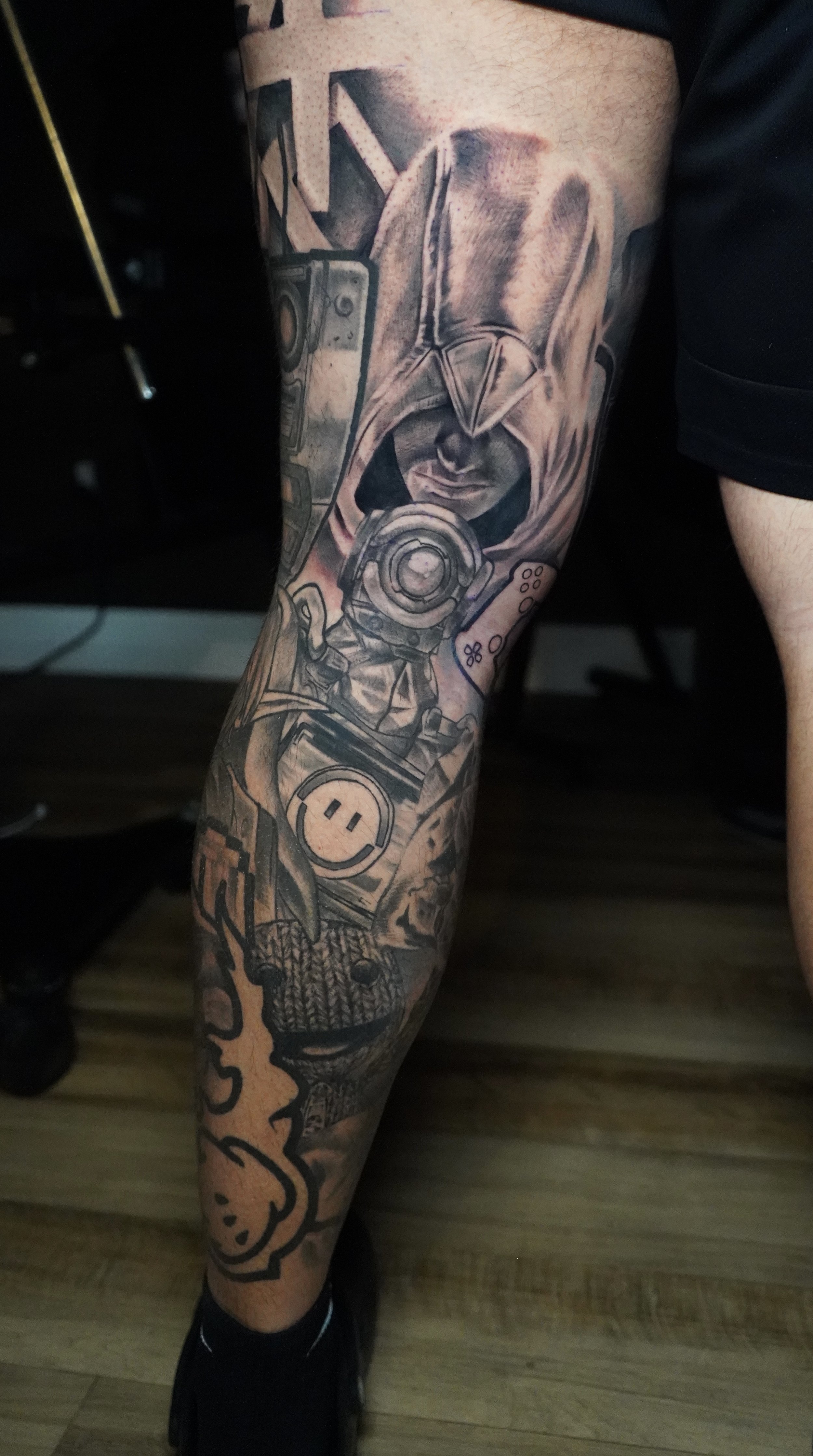 Tattoo of a hooded figure with a serious expression, surrounded by gaming controllers, a peace sign, and a smiling face, on a person's leg.