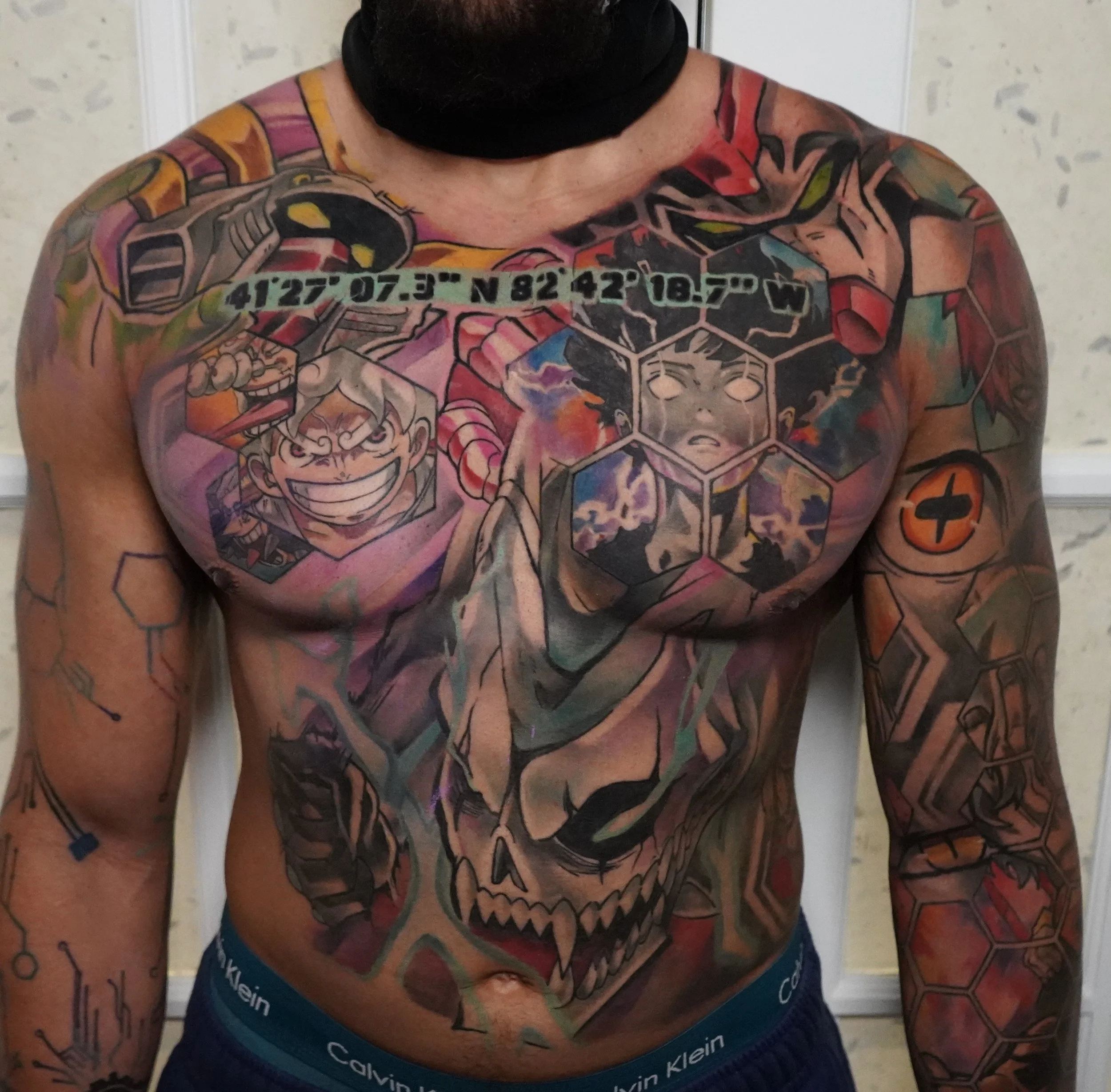 A man with a large, colorful tattoo covering his chest and arms, featuring a skull with geometric designs, anime characters, and scientific symbols.