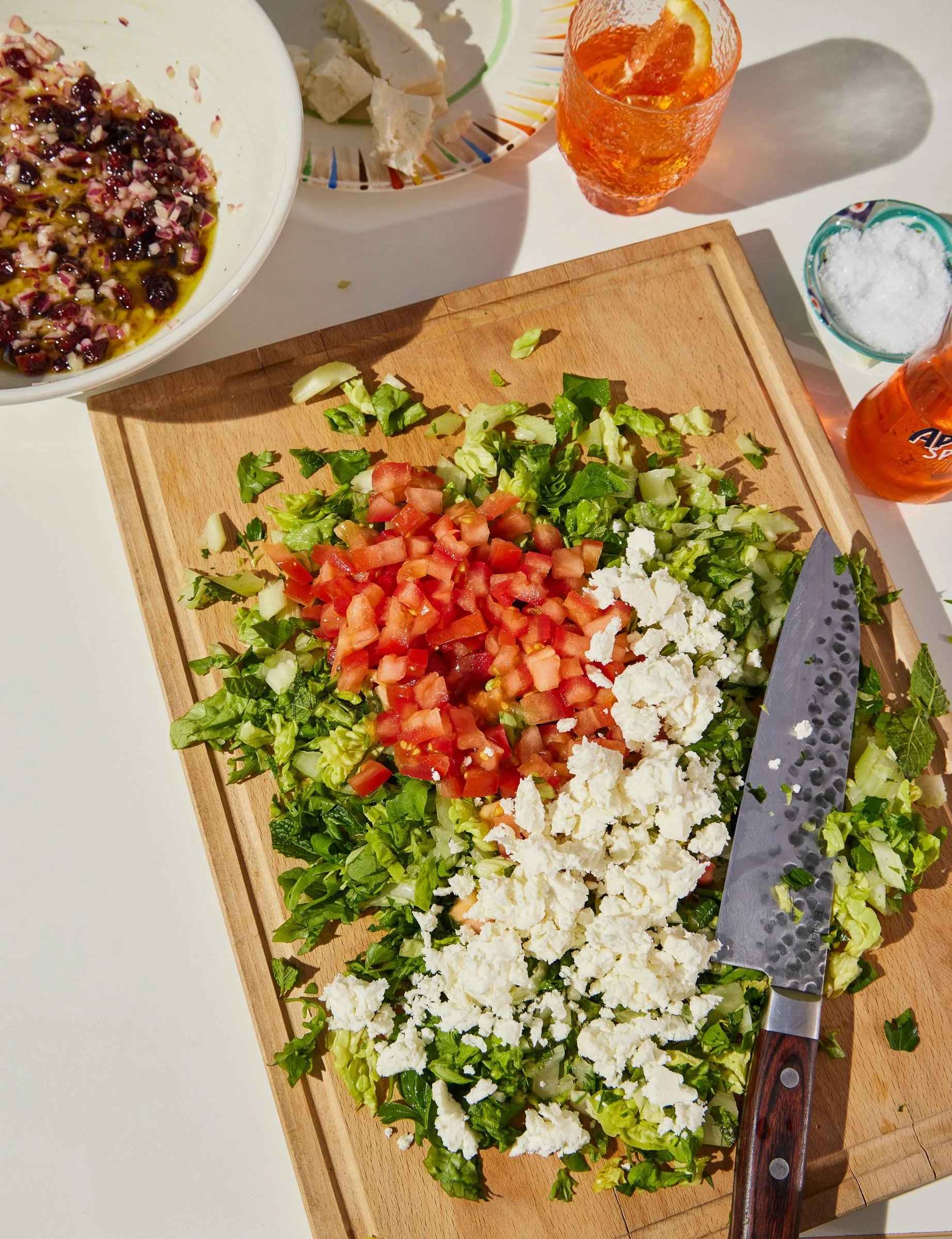 A wooden cutting board with chopped lettuce, diced tomatoes, crumbled feta cheese, and chopped cucumber, with a knife resting on it. In the background, there are plates with additional ingredients, orange drinks with ice, and a small bowl of salt.