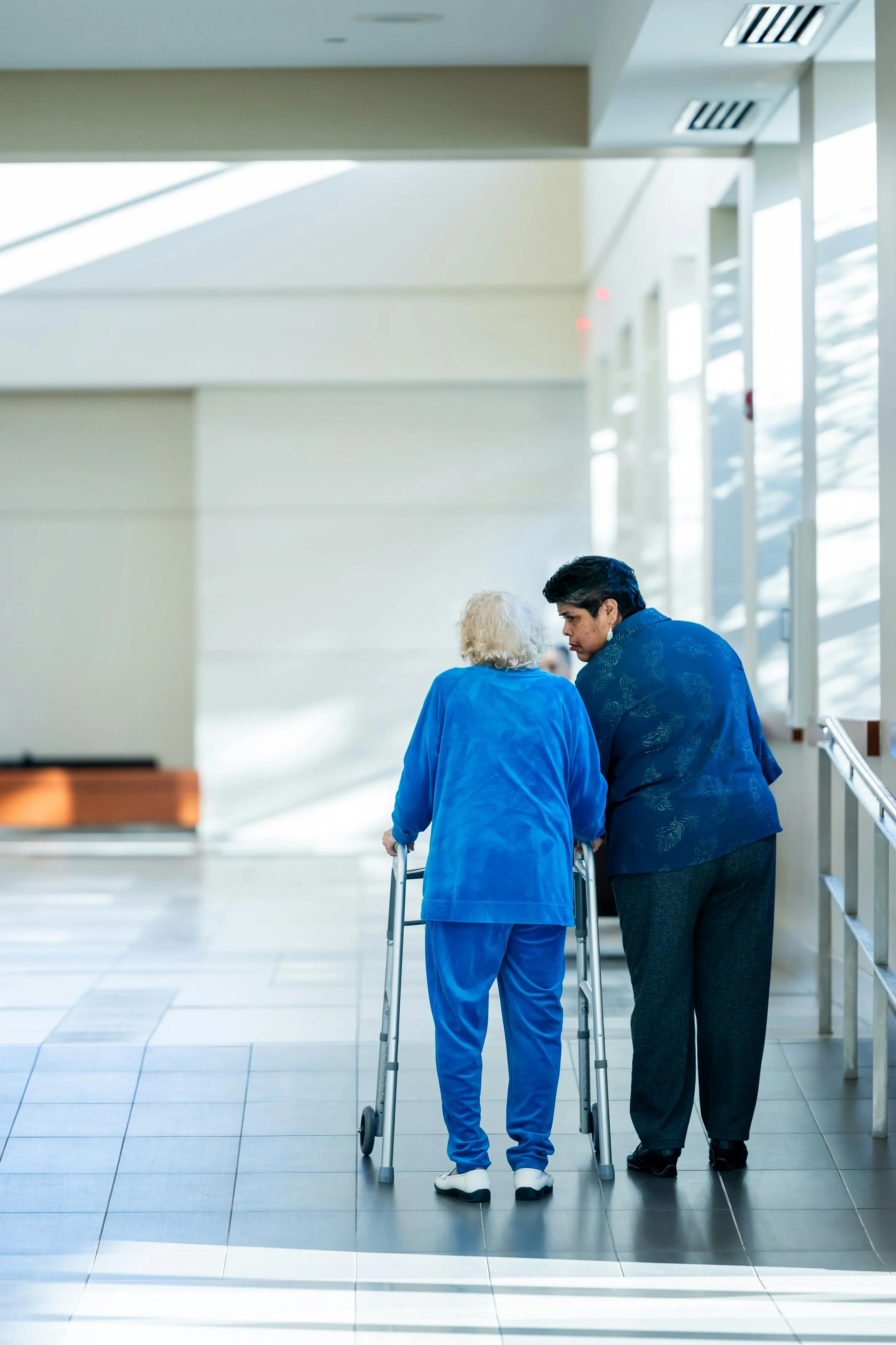 An elderly woman with white hair using a walker, accompanied by a man, walking through a brightly lit, modern hospital or healthcare facility corridor.
