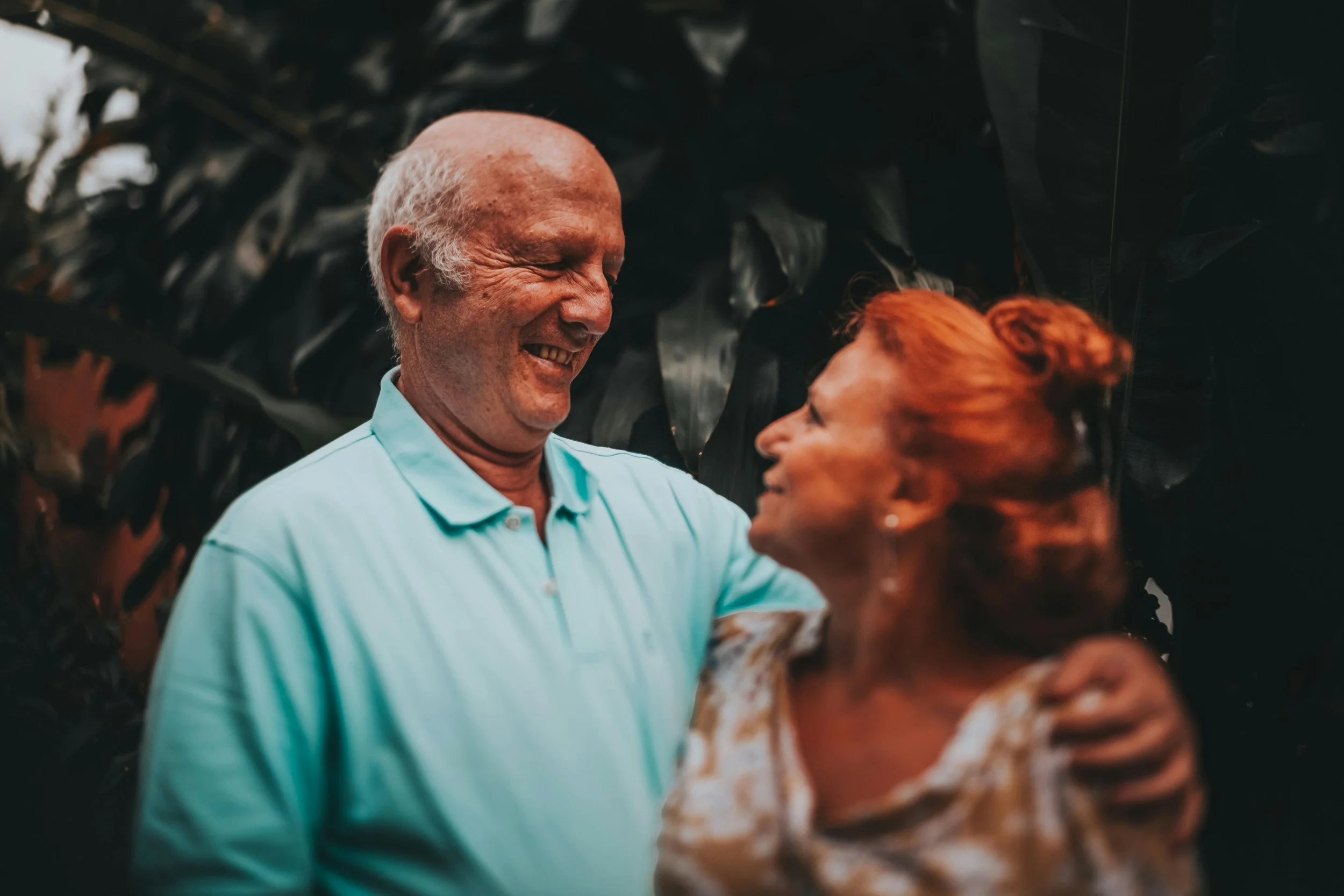A happy elderly man and woman sharing a joyful moment outdoors amidst dark green foliage, smiling at each other.