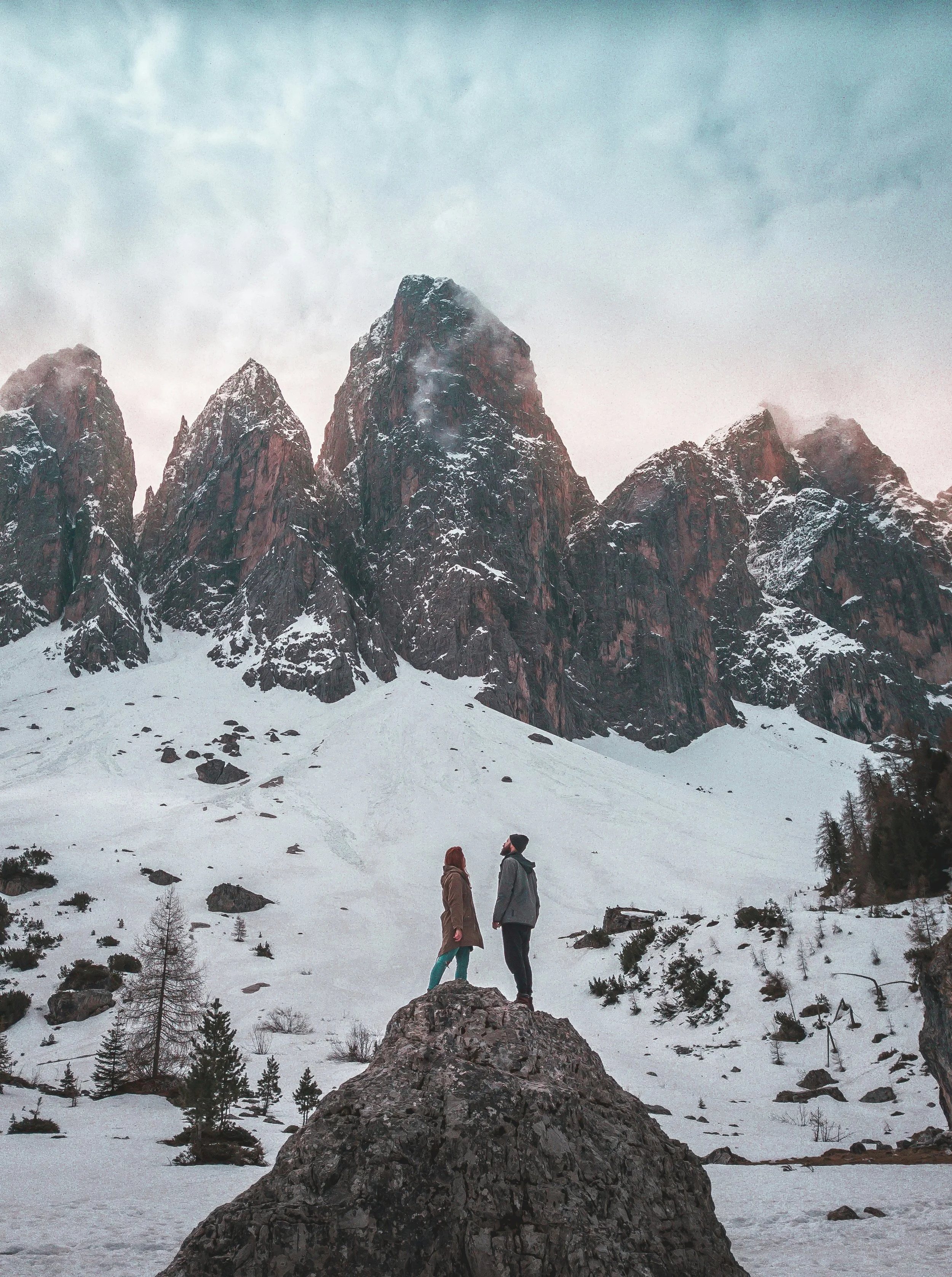 Two people standing on a large rock in a snow-covered landscape with tall mountains in the background.