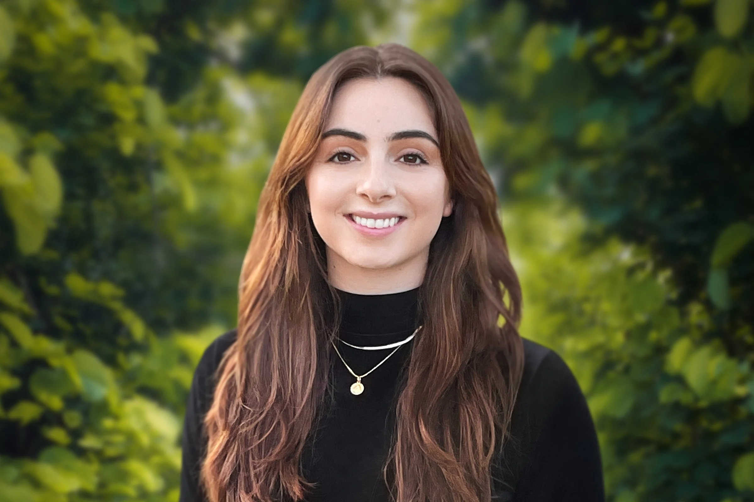 Young woman with long brown hair smiling in front of a green, leafy background.