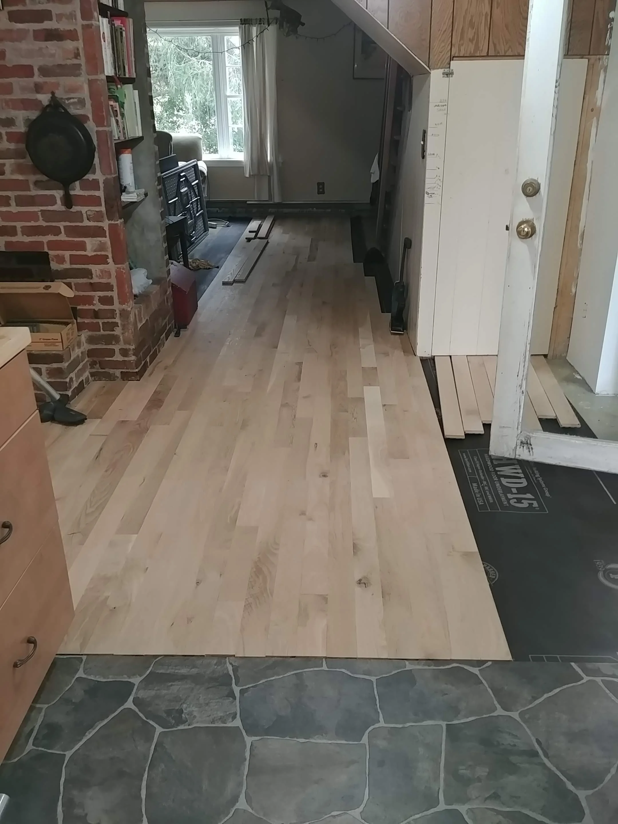 A room under renovation shows a transition from dark stone tile flooring in the foreground to light wood flooring in the background near a window. Construction materials and tools are visible, including a black workbench, wooden planks, and a partial