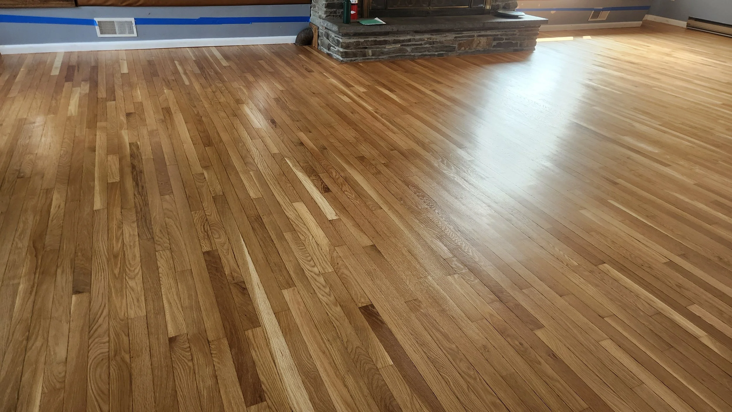Freshly refinished hardwood floor inside a house with a stone fireplace and a wall with blue and gray paint.