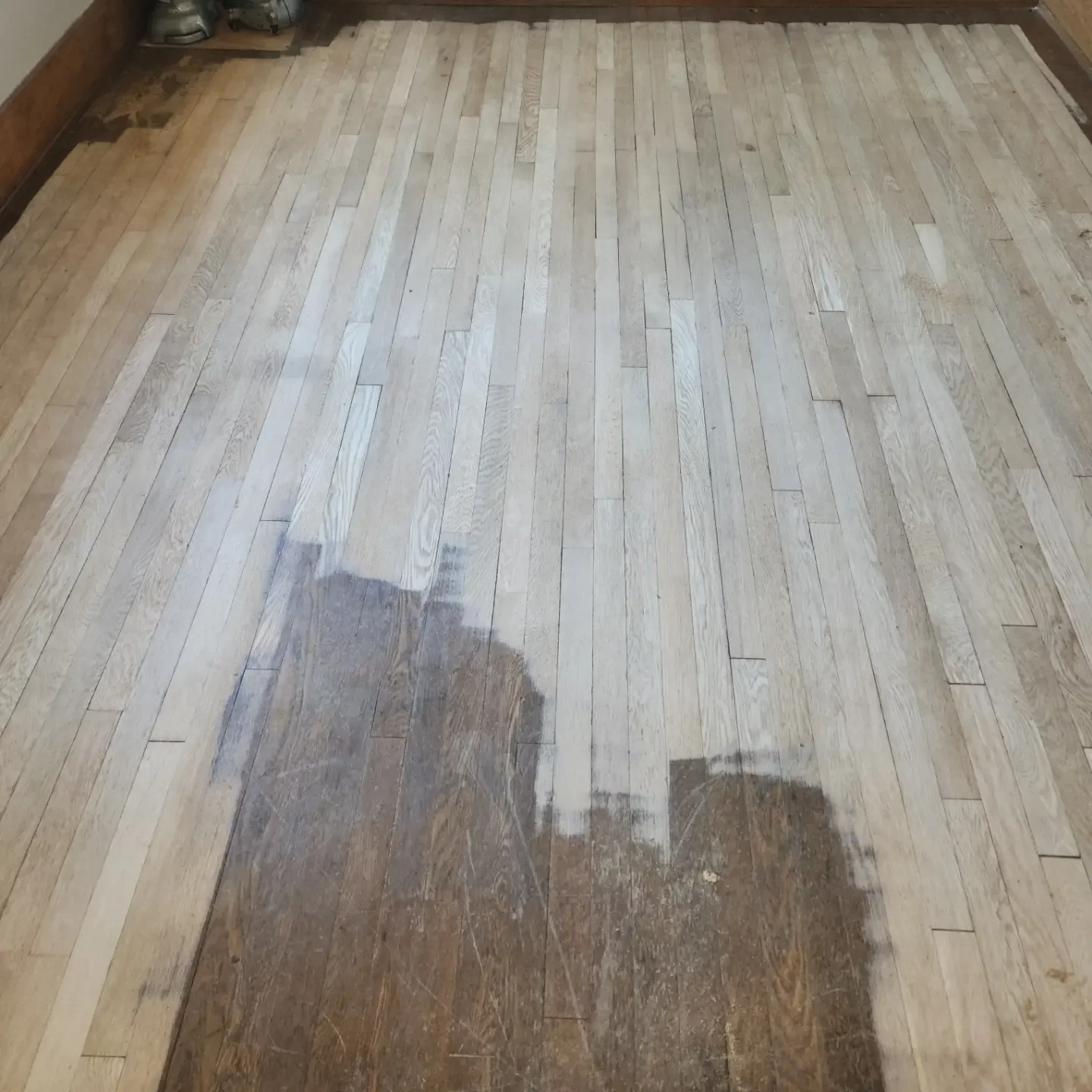 A hardwood floor in the process of refinishing, with a section shown as sanded and unfinished, contrasting with the older, darker, and more worn area nearby.