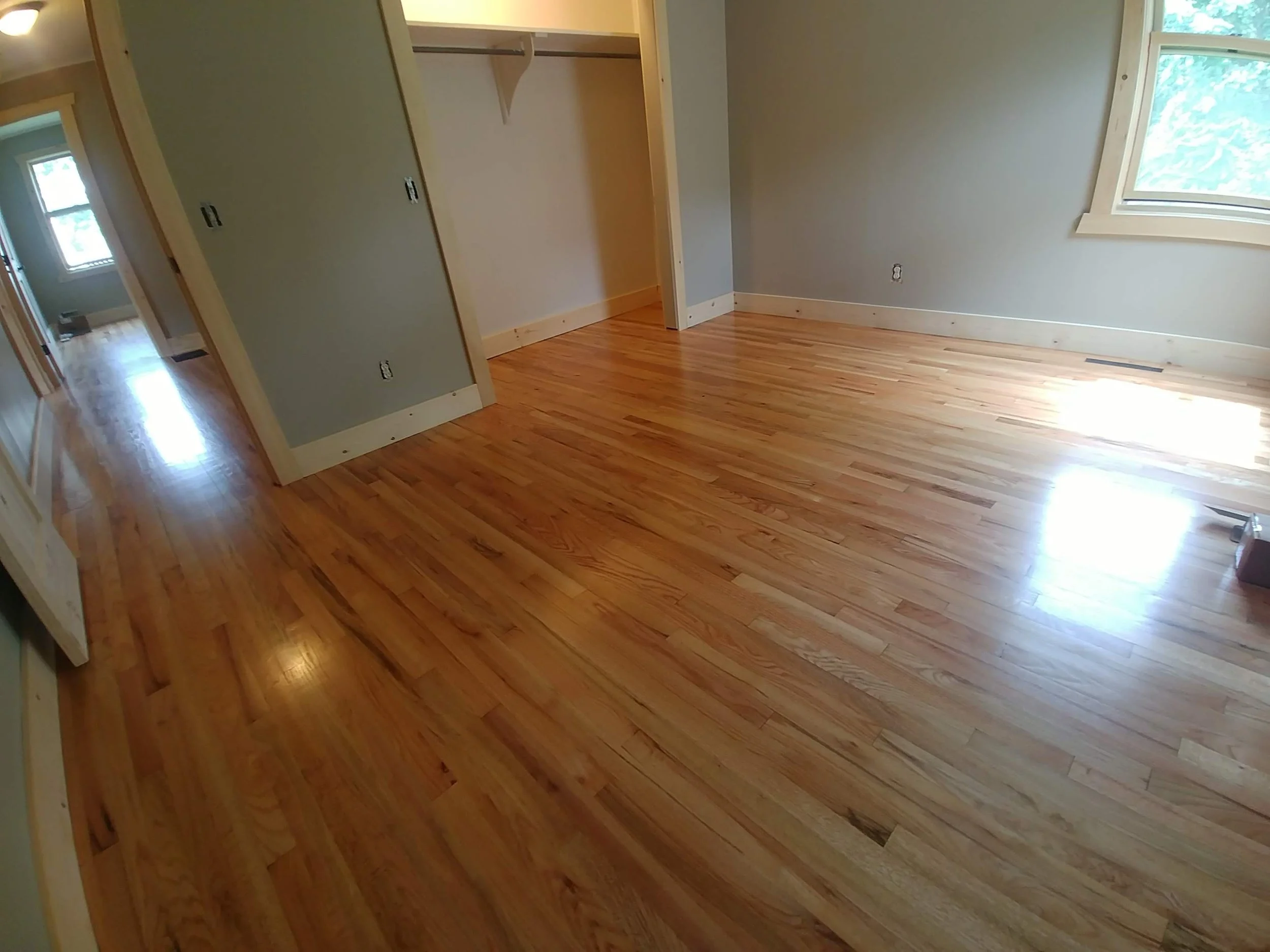 Empty room with hardwood flooring, a closet with sliding doors, a window, and a hallway visible in the background.