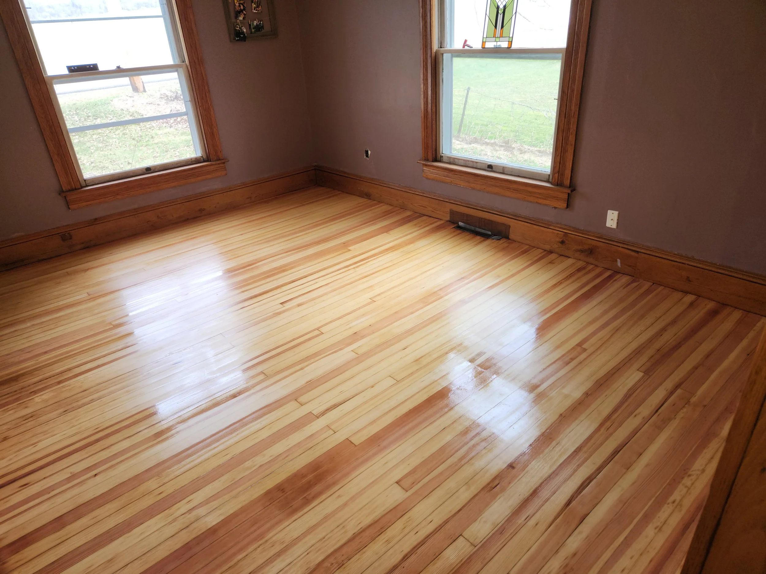 Empty room with hardwood floors, two windows, brown walls, and baseboards, with a view of a grassy outdoor area.
