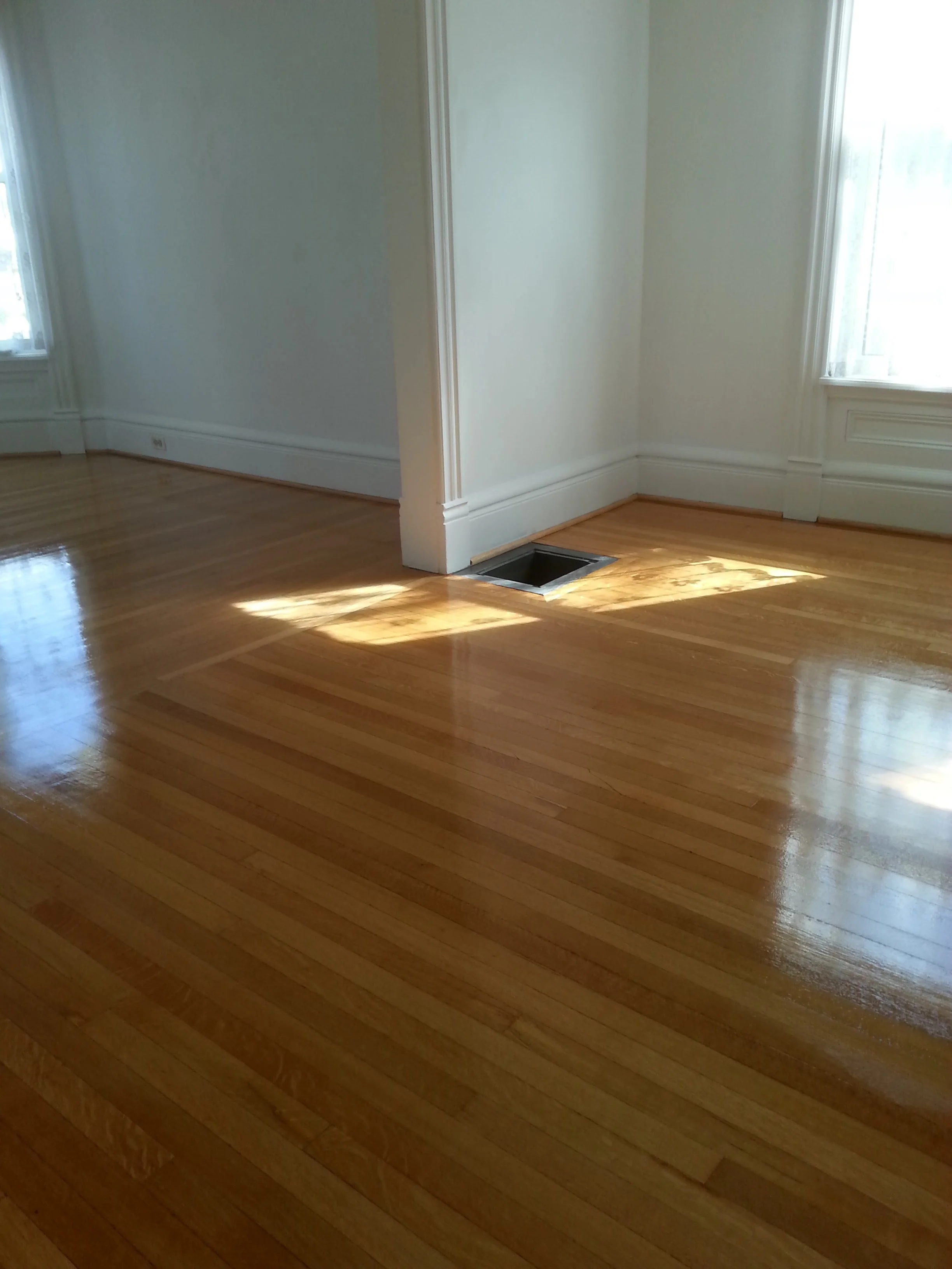 Empty room with hardwood floors, white walls, large windows, and a floor vent near the corner with sunlight shining through.