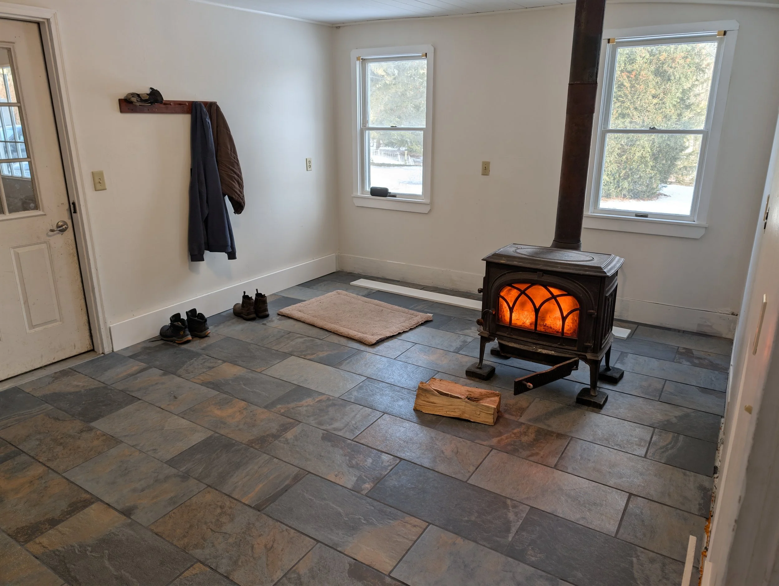 Room with basalt tile floors, two windows, a wood stove with a fire burning, a small beige rug, a pair of coats hanging on a wall-mounted shelf, two pairs of shoes on the floor, and a door on the left side.