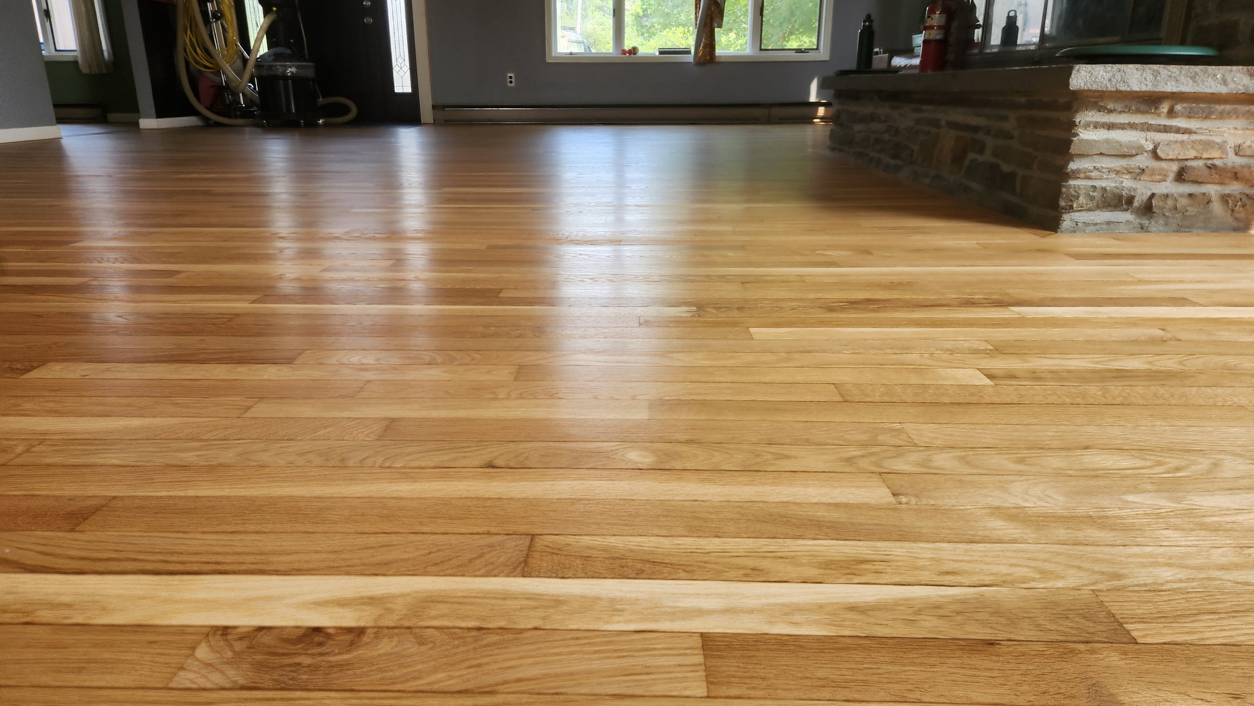 Photo of a polished wooden floor in a living room with a stone fireplace on the right and large windows in the background.