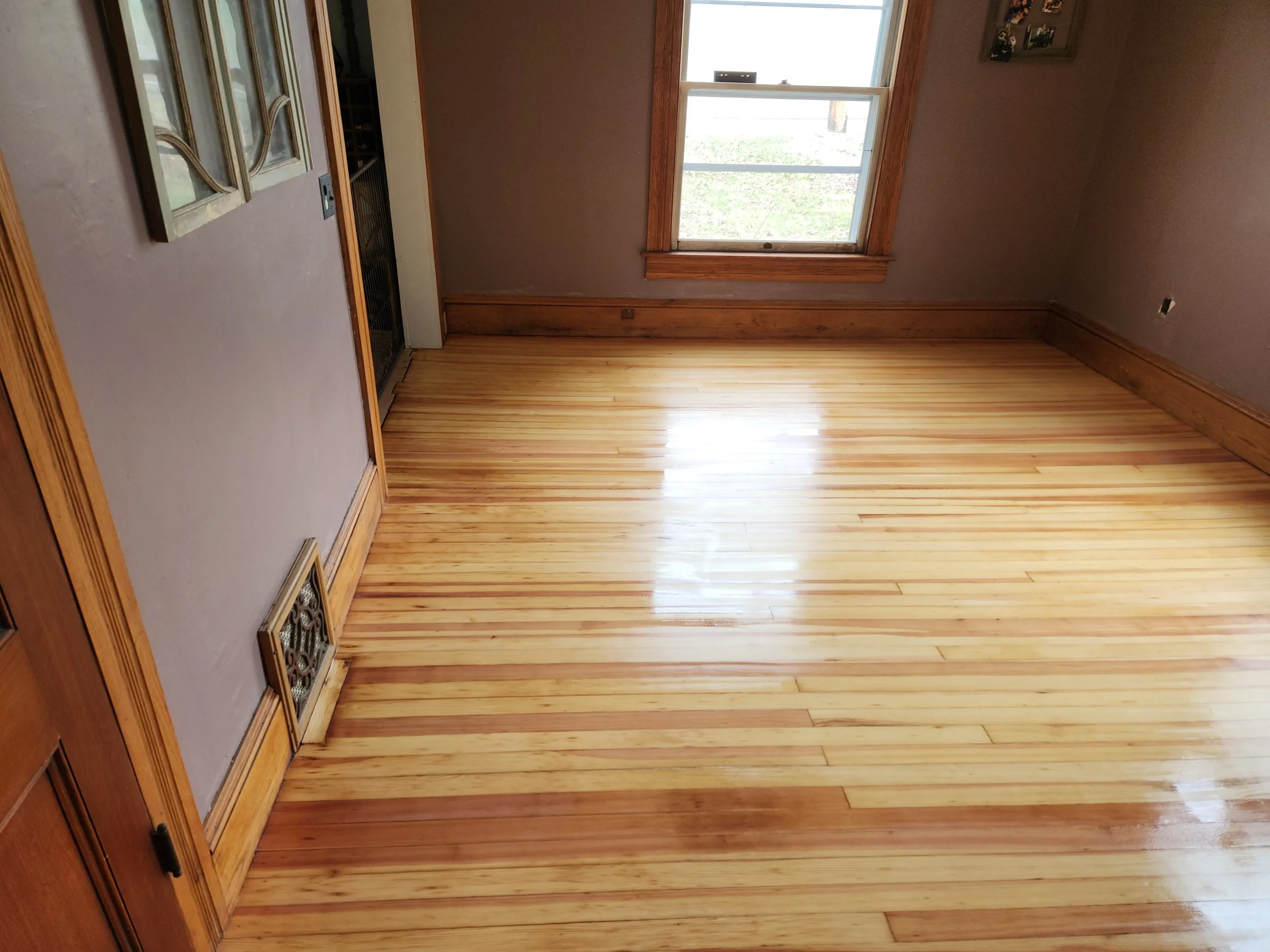 An empty room with new wooden flooring, a window with wooden trim, and a painted wall with a picture frame. There's a vent on the floor near the corner, and the room is illuminated by natural daylight.