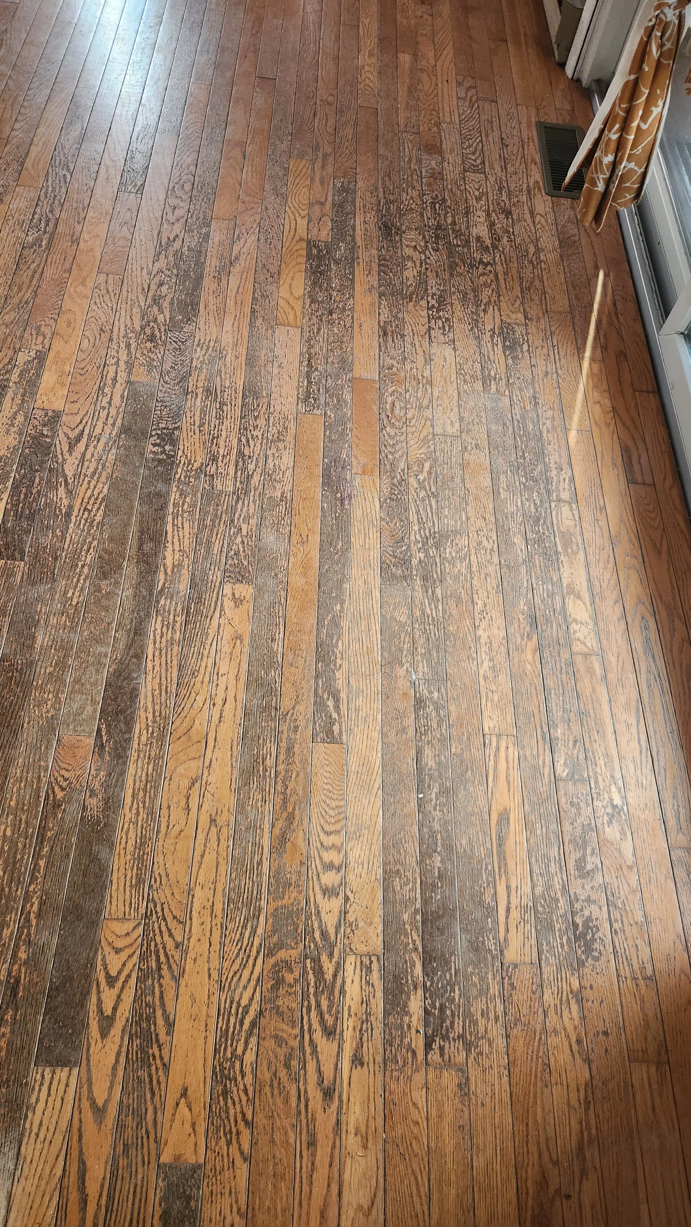 Wooden floor with noticeable wear, scratches, and discoloration near the edges of a room.