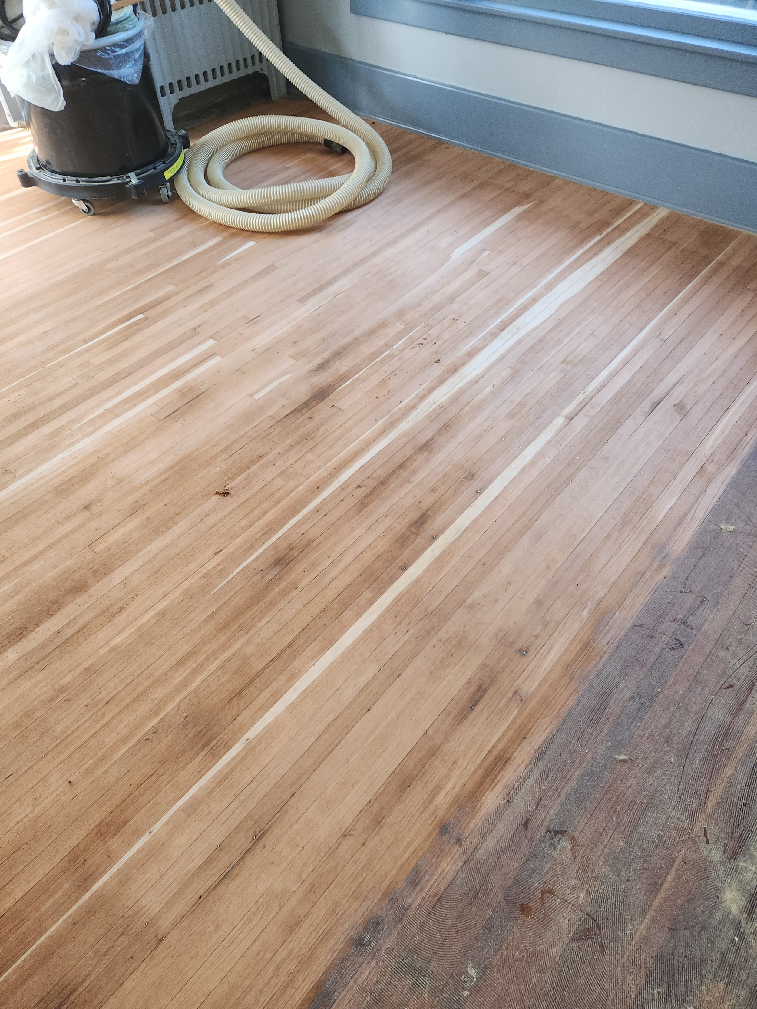 A wooden floor being refinished with a machine, with a vacuum and a hose nearby, near a wall with a gray protection board.