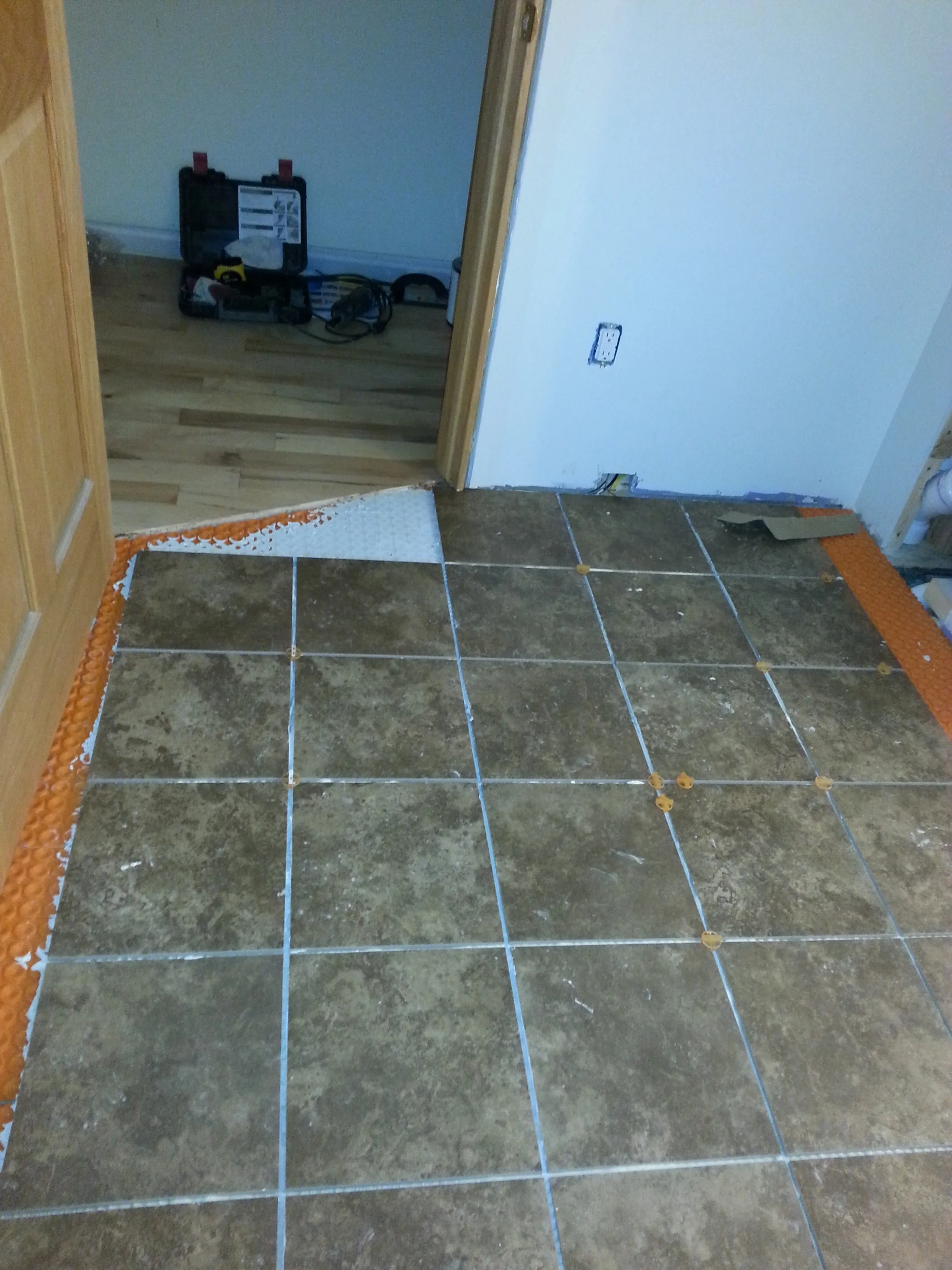 Undertile heating system installation with heating mats and mesh, partially installed on a tiled kitchen floor, with a doorway leading to a room with hardwood flooring.