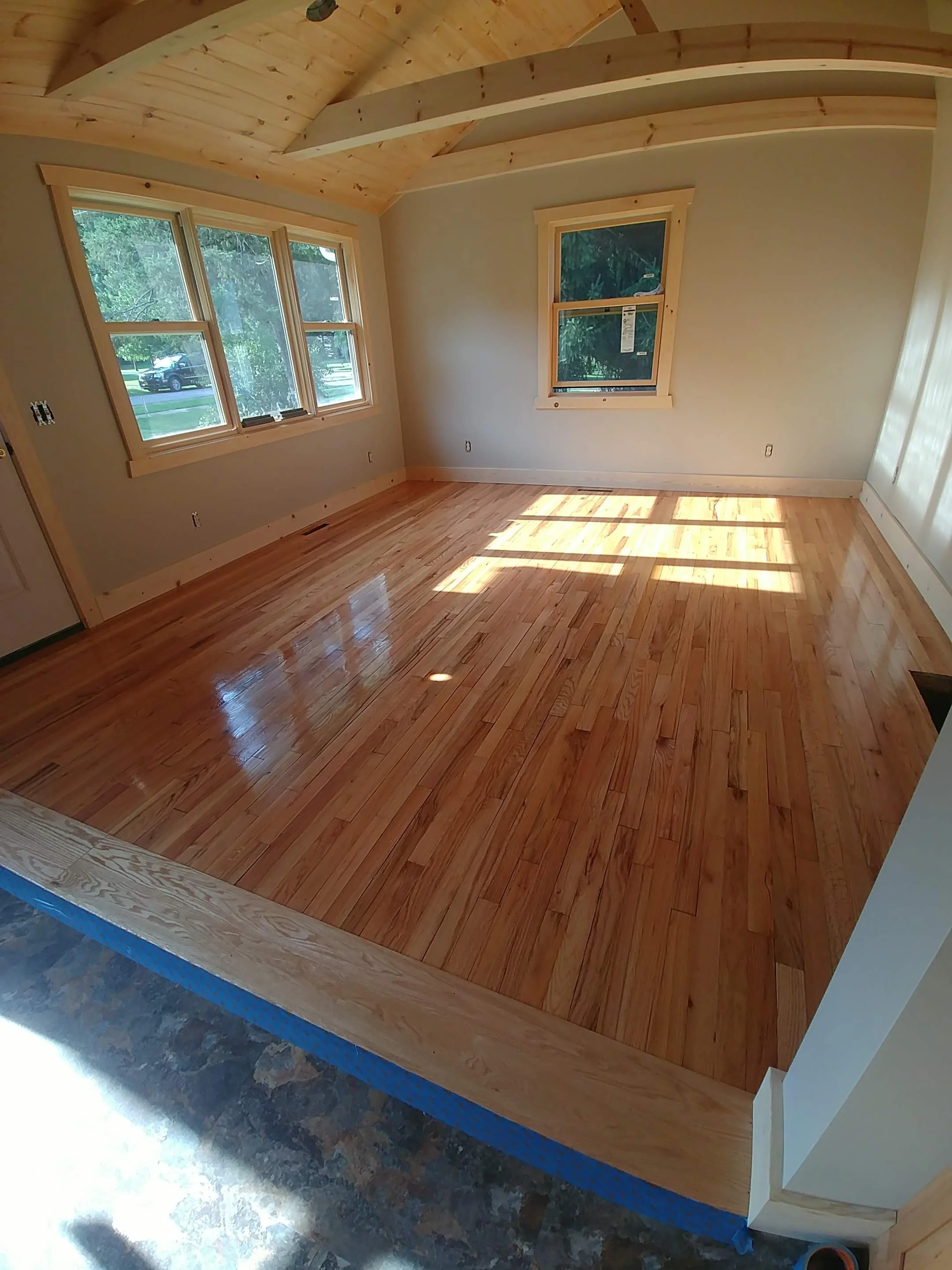 Empty room with wooden floors, light-colored walls, and large windows letting in natural light, with a wooden ceiling and trim around the windows.