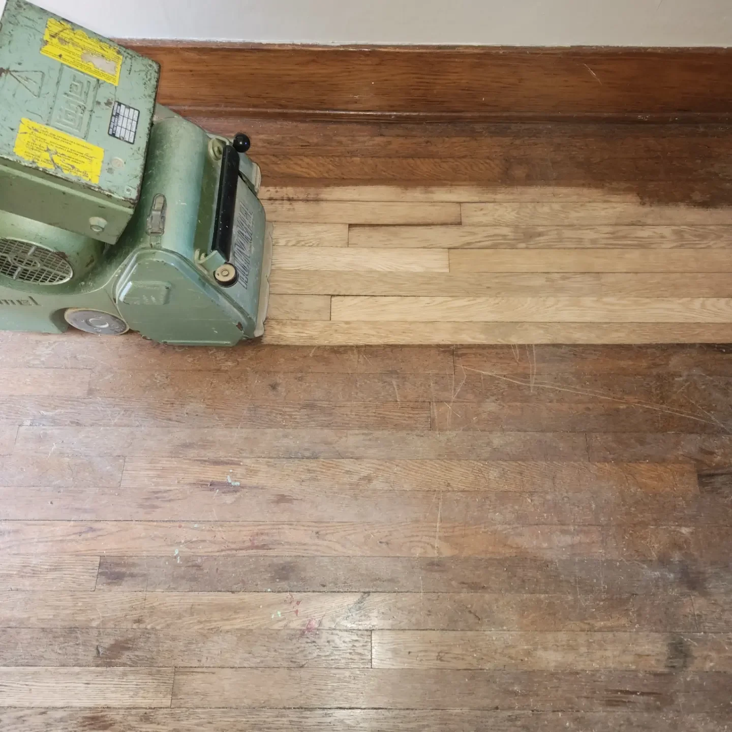 A section of hardwood flooring in a room, showing a transition from a darker wood finish to a lighter wood section. An industrial green machine is partially visible in the top left corner.
