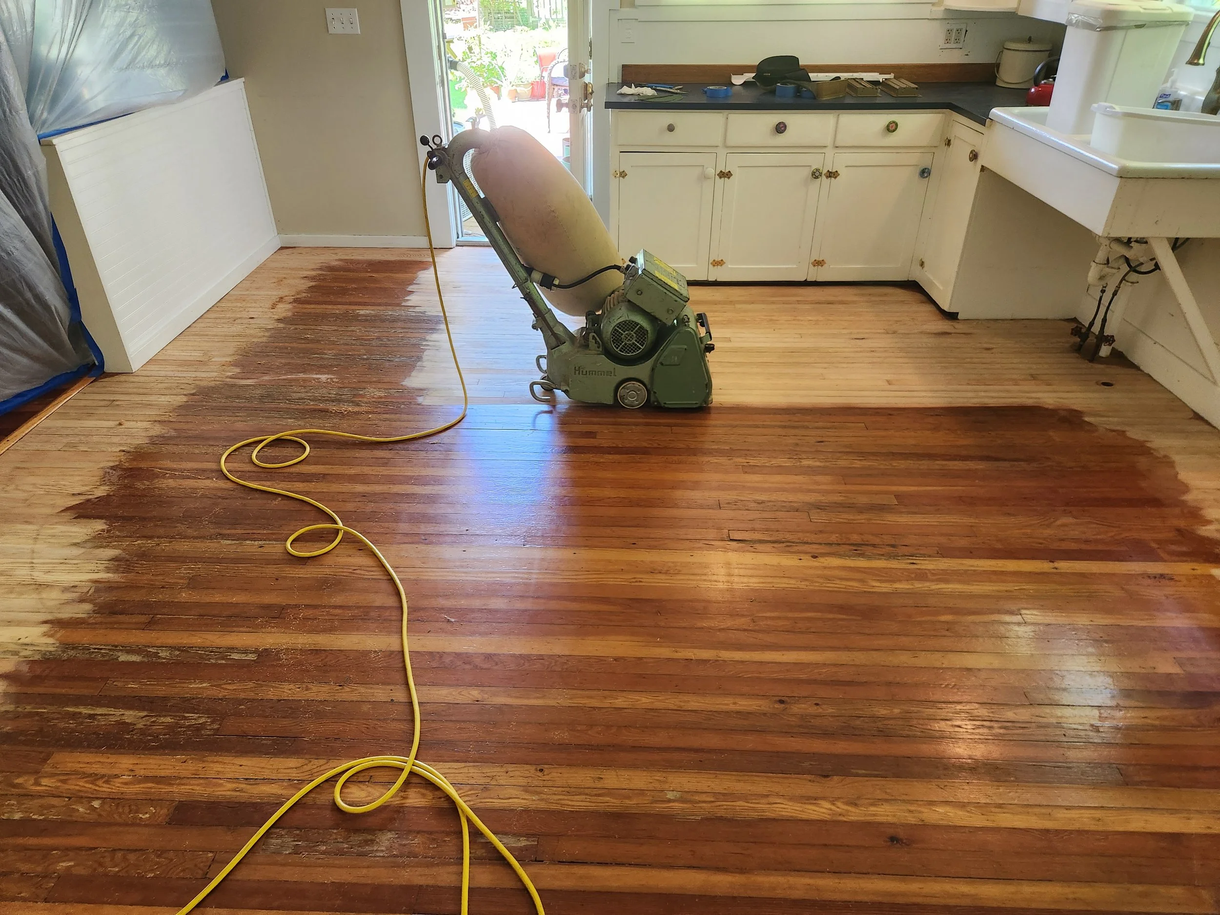 A hardwood floor being sanded and refinished with a floor sander in a kitchen, with vacuum hose and electrical cord visible.